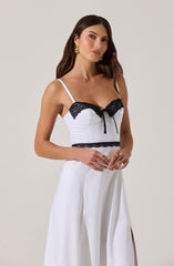 A woman wearing a white sleeveless jumpsuit with thin spaghetti straps. The top has black lace trim along the neckline and a small black bow at the center. The waistline features a black lace band. The jumpsuit has wide-leg pants with pleats and a side slit. The woman has long brown hair, gold earrings, and gold rings on her fingers.