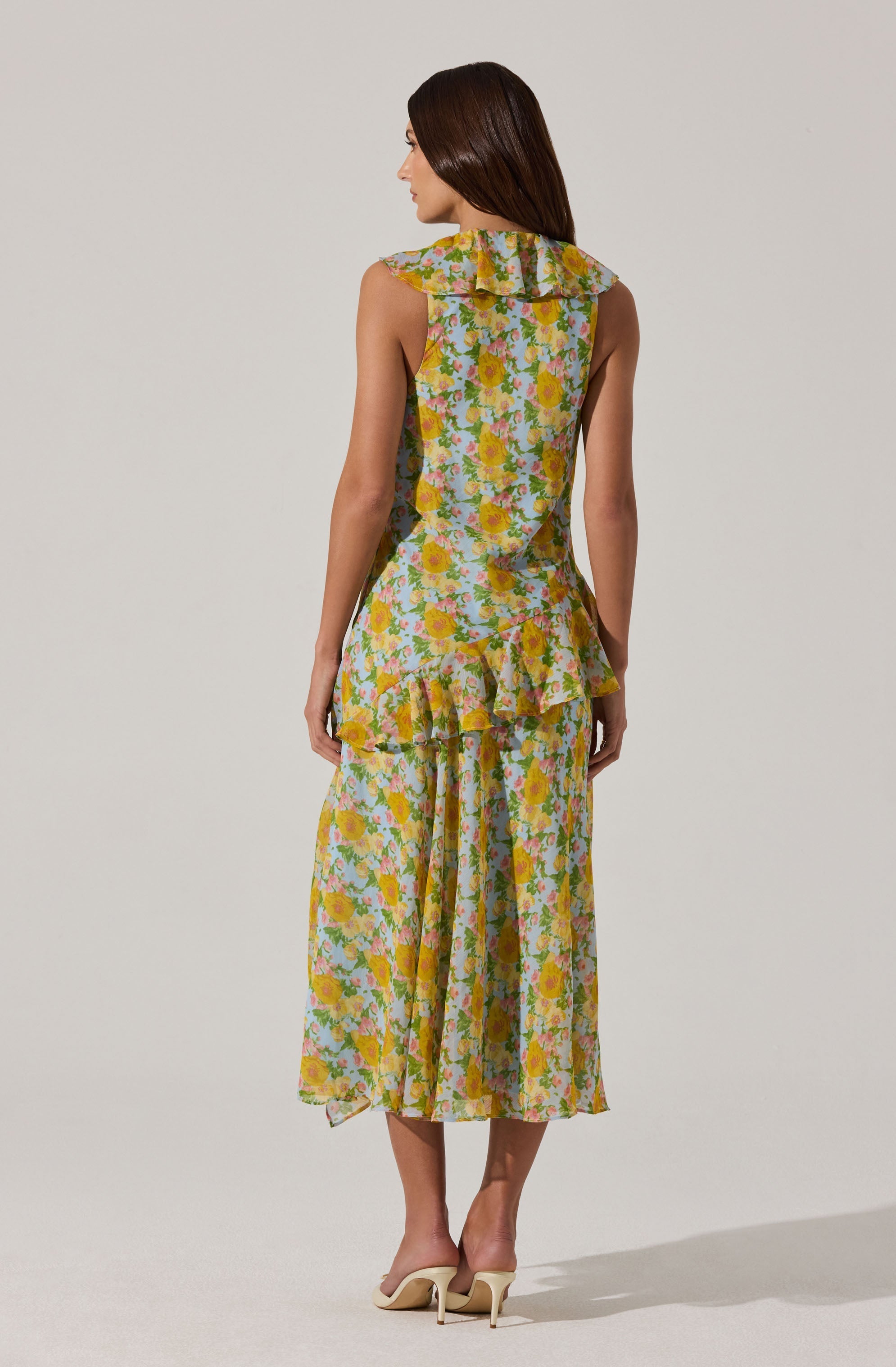 A woman is wearing a sleeveless, floral dress with a ruffled collar and a ruffle detail around the waist. The dress features a colorful print of yellow, pink, and blue flowers on a light background, and it has a flowy, midi-length skirt. She is also wearing beige high-heeled shoes.