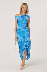 A woman is wearing a blue tie-dye dress with an asymmetrical off-the-shoulder neckline, ruched details around the waist, and a ruffled, high-low hemline. She is holding a small white woven handbag and wearing white open-toe high heels.