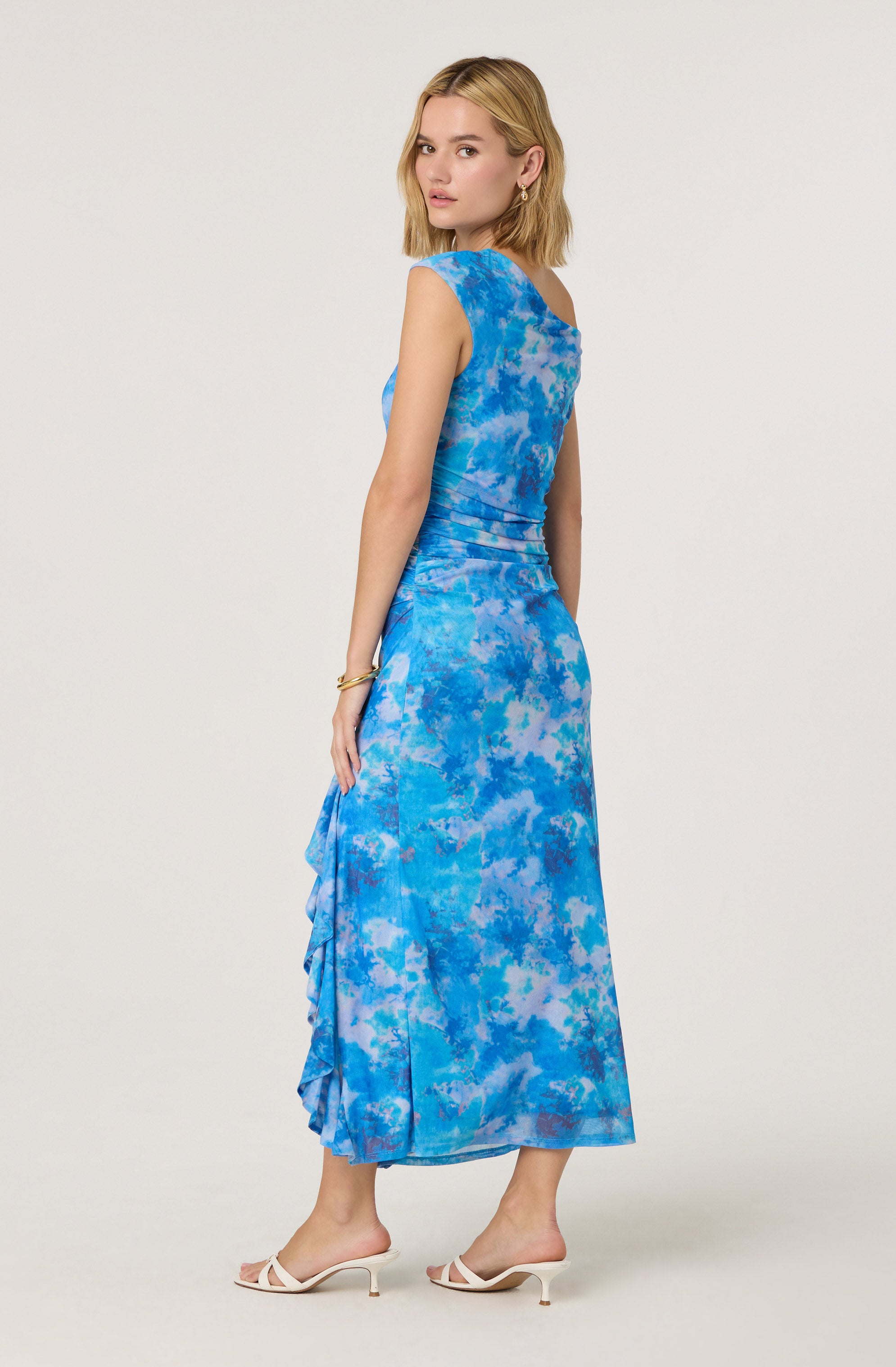 A woman is wearing a sleeveless, ankle-length dress with a blue and light blue abstract tie-dye pattern. The dress has a fitted waist, a slightly draped ruffle detail on one side, and a round neckline. She is accessorizing with gold hoop earrings and a bracelet, and is wearing white open-toe heeled sandals.
