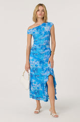 A woman is wearing a one-shoulder dress with a blue tie-dye pattern. The dress has a fitted waist and a ruffled asymmetrical hem that features a slit. She accessorizes with a gold bracelet, white open-toe heels, and carries a small white handbag.