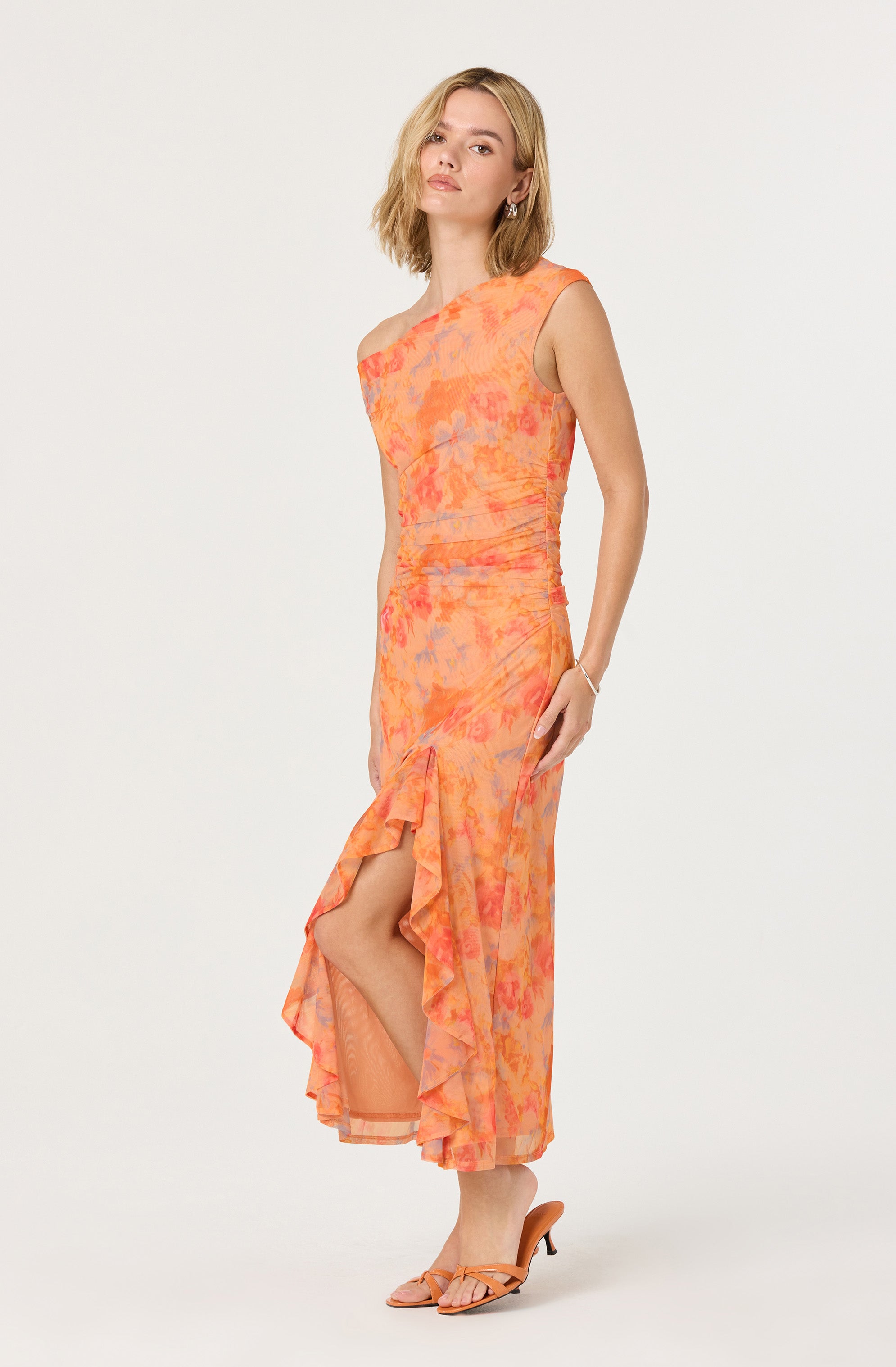 The dress is sleeveless with an asymmetrical neckline and features a floral pattern in shades of orange, pink, and purple. It has a fitted bodice and a flowing, ruffled skirt with a slit on one side, revealing part of the leg. The fabric appears lightweight and slightly sheer. The dress is paired with matching peach-colored heels.