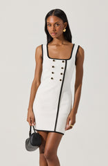 A woman is wearing a sleeveless, fitted white dress with black trim along the neckline, straps, and a vertical line down the front. The dress features two rows of gold buttons on the front. She accessorizes with gold earrings and a gold ring, and holds a small black handbag in her right hand.
