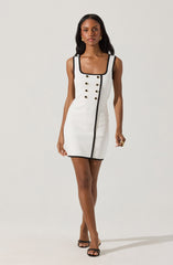 A woman is wearing a sleeveless, fitted white dress with black trim along the square neckline, armholes, and a vertical line running from the neckline to the hem. The dress features two rows of gold buttons on the front, arranged in a double-breasted style. She completes her outfit with black open-toe high-heeled sandals and gold hoop earrings.