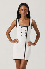 A woman is wearing a fitted sleeveless dress that is primarily white with black trim along the neckline, armholes, and a vertical line running down the front. The dress features decorative black buttons arranged in two vertical rows on the front. The dress has a square neckline and a straight hemline that ends above the knees. She accessorizes with gold earrings and rings.