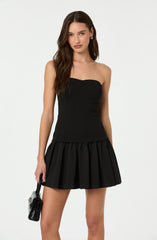 A woman is wearing a strapless black mini dress with a fitted bodice and a flared, pleated skirt. She is holding a small black handbag in her right hand.
