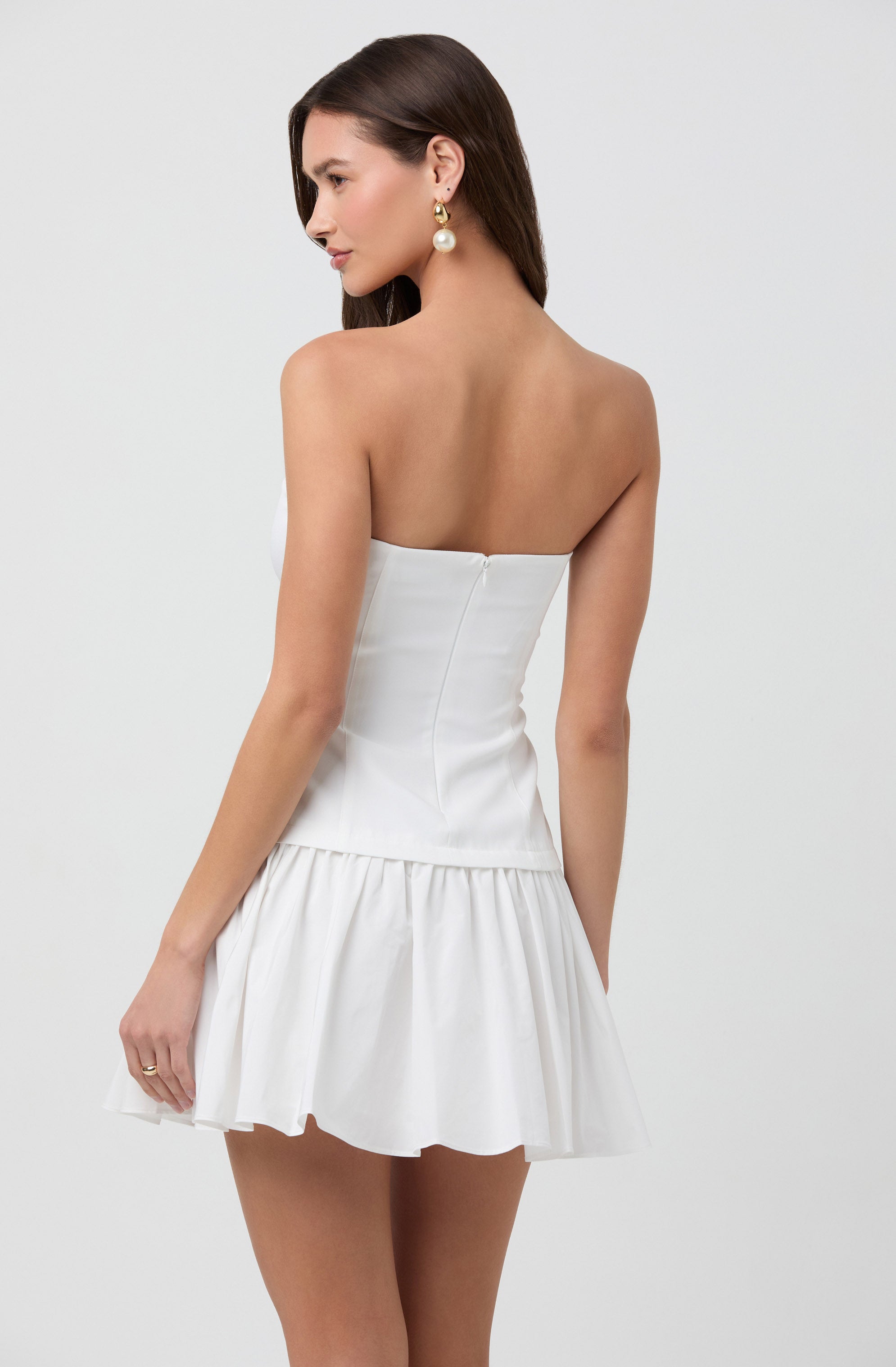 A woman is wearing a strapless white dress with a fitted bodice and a ruffled, flared skirt. She is accessorized with long, dangling earrings featuring a large pearl and a smaller decorative element.