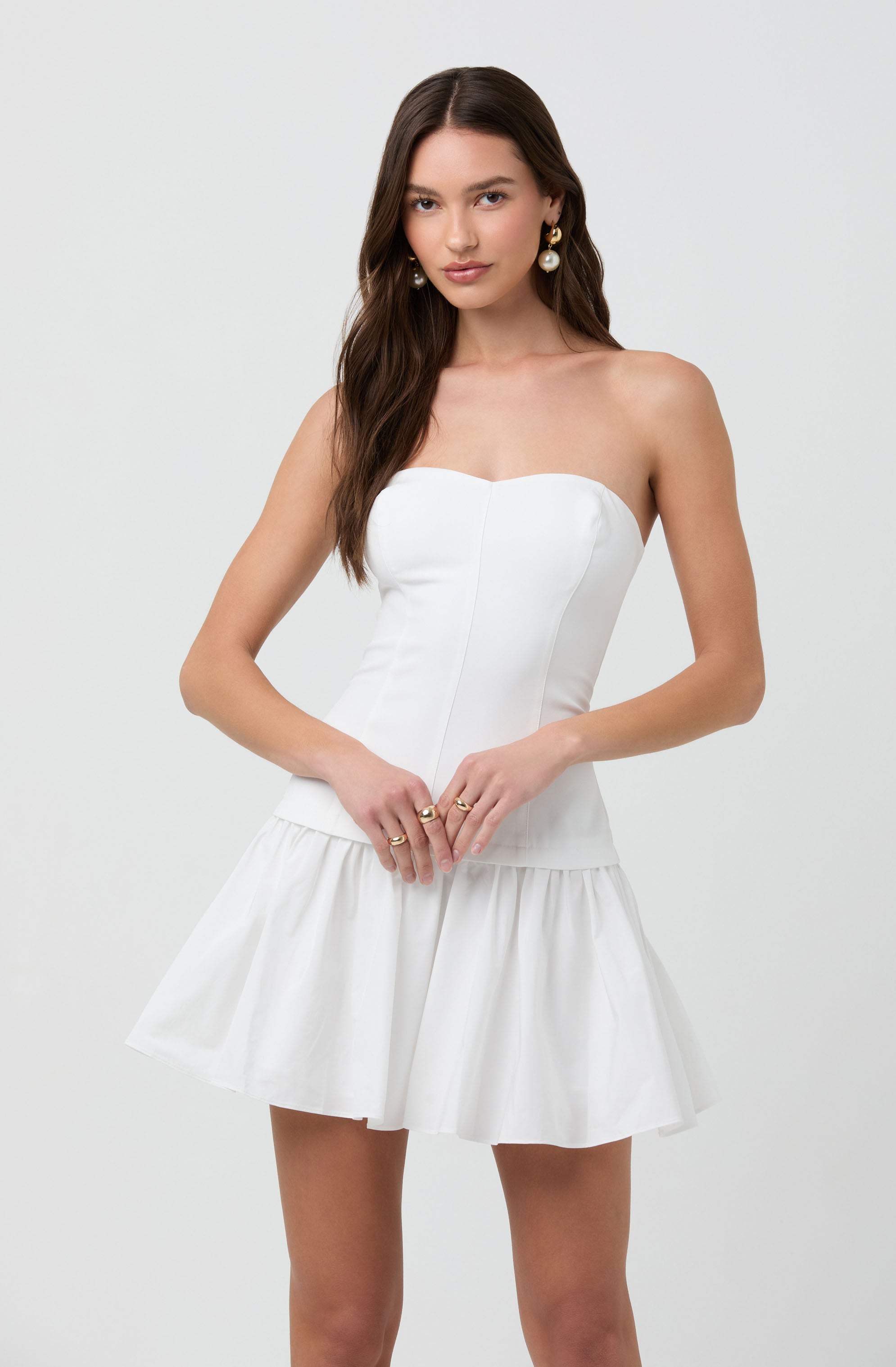 A woman is wearing a strapless white dress with a fitted bodice and a flared, ruffled skirt. She is accessorized with gold earrings and rings.