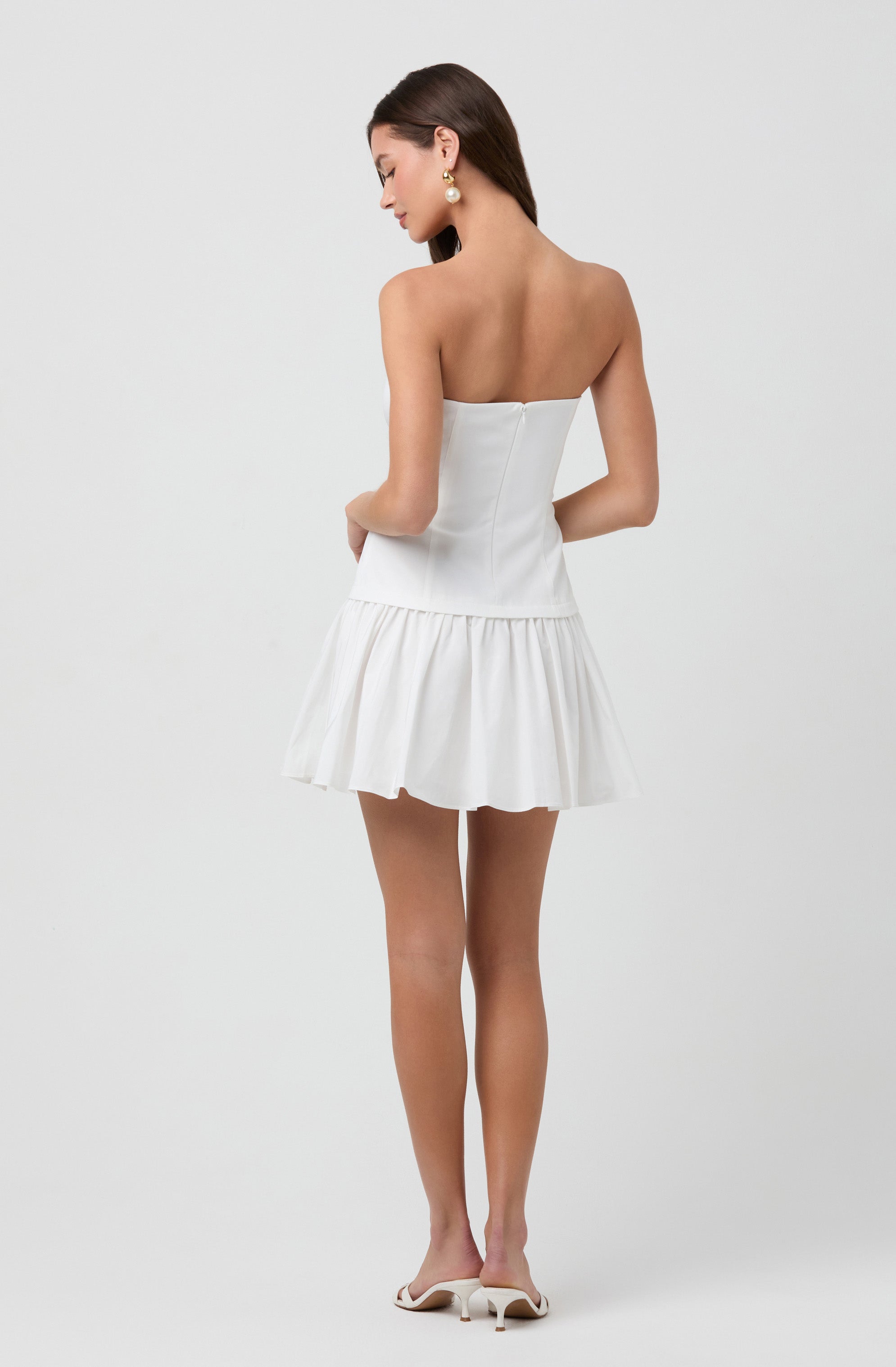 A woman is wearing a strapless white dress with a fitted bodice and a gathered, ruffled skirt that falls above the knees. She has large pearl earrings and is wearing white high-heeled shoes.