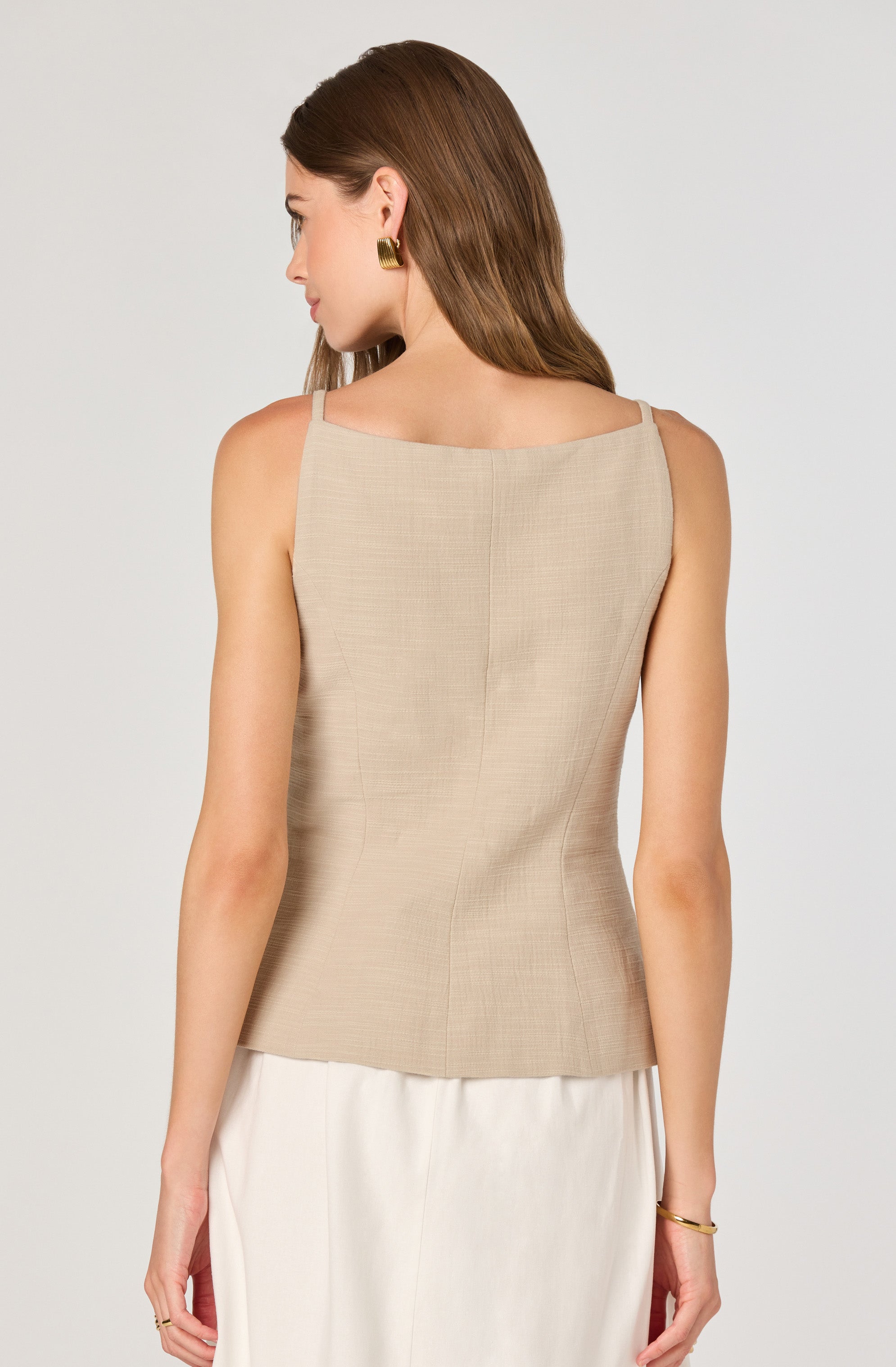 A woman is wearing a sleeveless, beige top with thin straps and a subtle texture. The top has a simple, classic cut with a straight neckline and vertical seam details running down the back. She pairs it with creamy white pants. She accessorizes with gold earrings and a gold bracelet.
