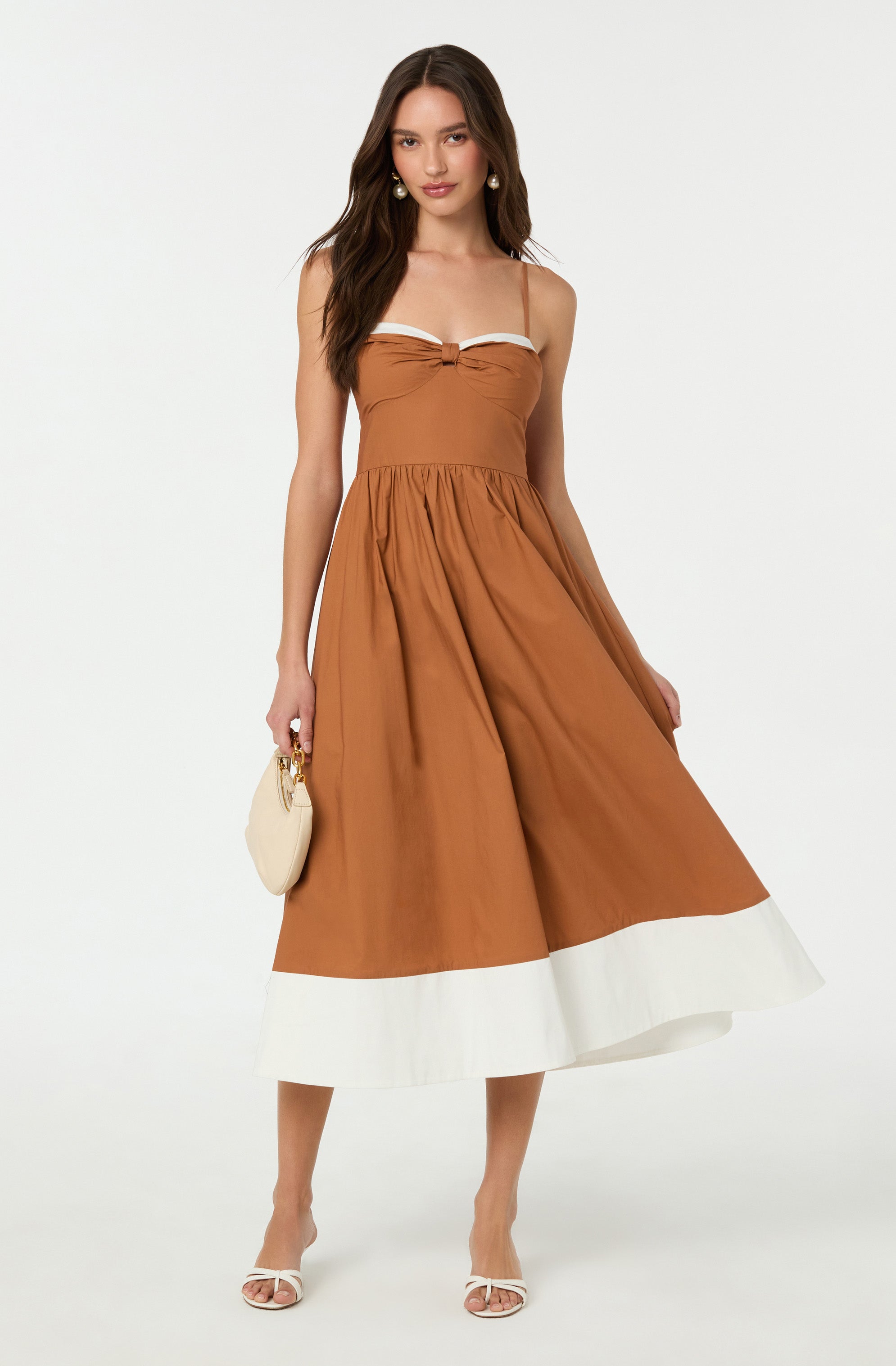 A woman is wearing a sleeveless, brown midi dress with thin straps and a decorative bow at the center of the chest. The dress has a gathered waist and a wide white band at the hem. She is also wearing white open-toe heels and holding a small, curved cream-colored purse with a gold handle.
