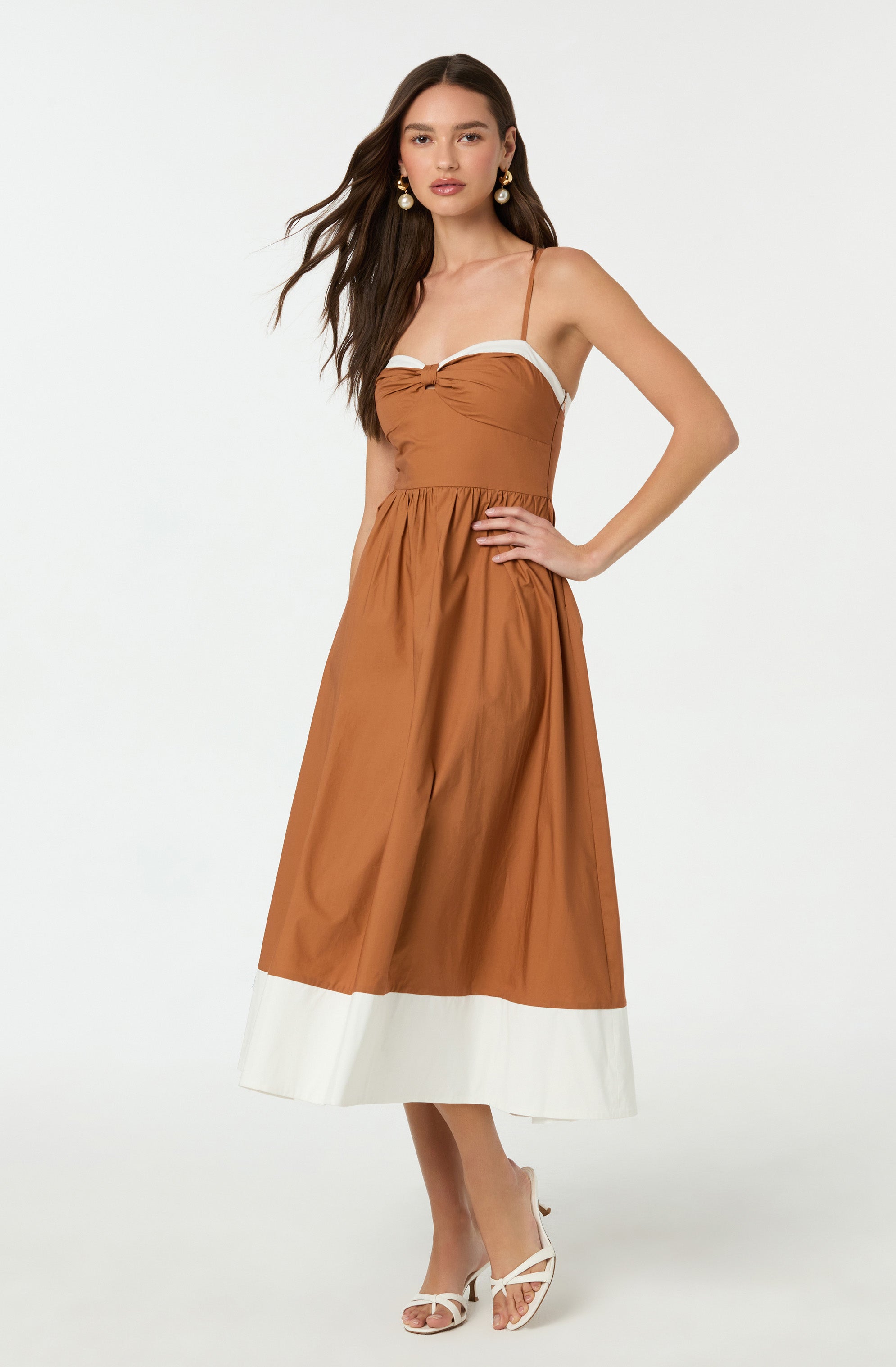 A woman is wearing a sleeveless, brown midi dress with thin spaghetti straps and a white trim along the top edge. The dress has an elastic waistband and a white band at the hem. She is accessorized with large white and gold earrings and is wearing white high-heeled sandals.