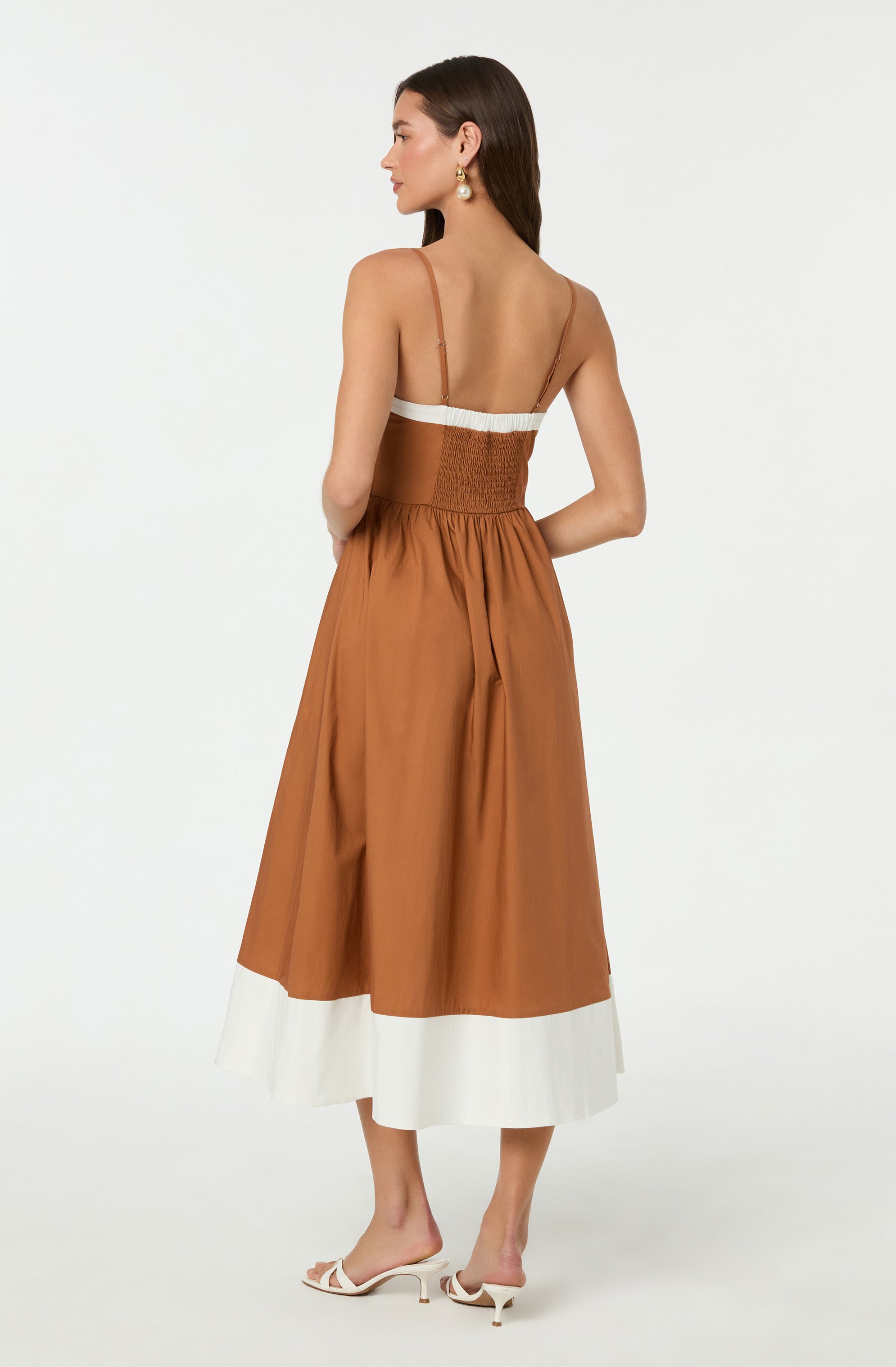 A woman is wearing a sleeveless, strappy dress with a fitted bodice and a full, ankle-length skirt. The dress is primarily a warm caramel brown with a wide, white band at the hem. She is also wearing white, open-toed high-heeled sandals.