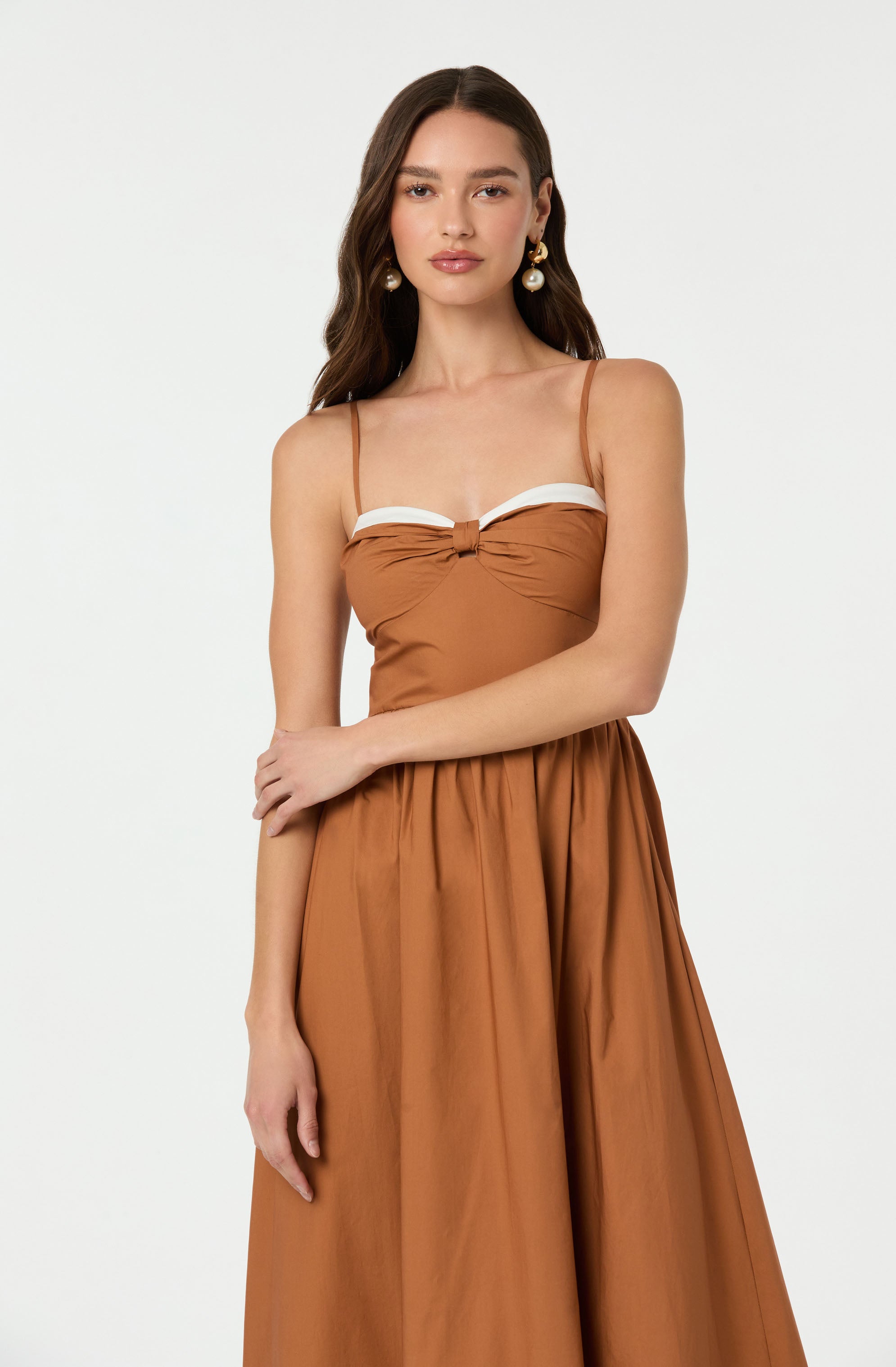 A young woman is wearing a burnt orange dress with spaghetti straps. The top of the dress features a white band detail across the bust, with a small knot in the center. The skirt is flowy and gathered at the waist, creating a full, elegant silhouette.