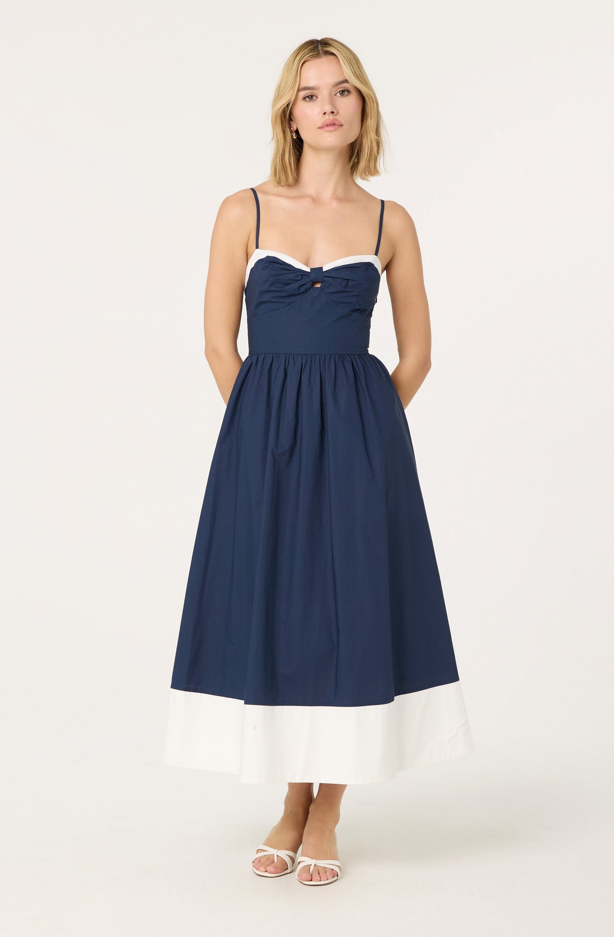 A woman is wearing a sleeveless, navy blue maxi dress with thin spaghetti straps. The dress features white trim along the top edge and a small bow detail at the center of the chest. The skirt is gathered at the waist and has a wide white band at the hem. She is also wearing white open-toe heels.