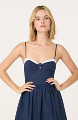 A woman is wearing a sleeveless, navy blue dress with thin spaghetti straps. The dress features a large bow detail at the bust with a small cut-out below the bow, and a white trim along the top edge. The skirt of the dress is gathered and falls loosely.