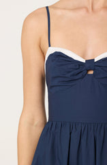 A person is wearing a sleeveless dress with thin straps. The dress has a navy blue bodice with a gathered, ruched detail in the center that is tied into a knot. The top edges of the dress are lined with a contrasting white fabric. The skirt portion of the dress is also navy blue with a gathered, flowing design.
