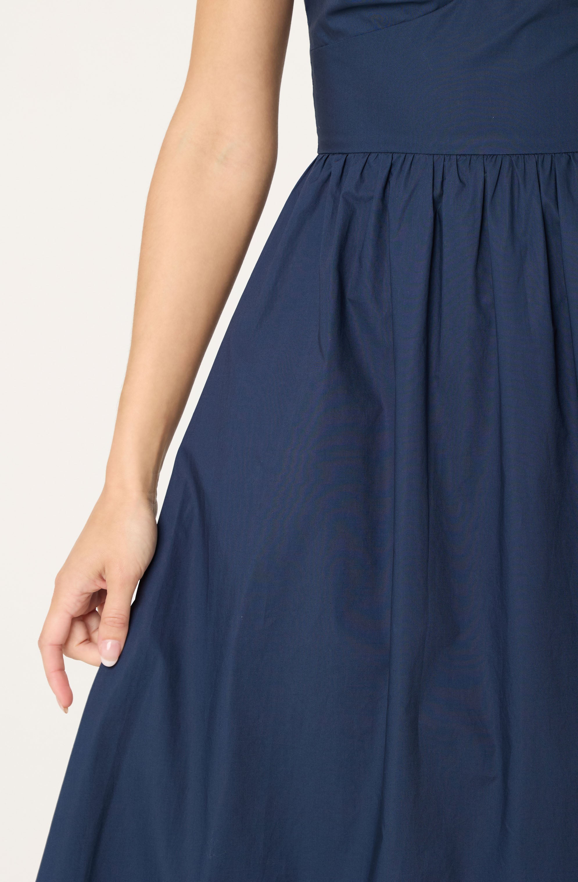 A person is wearing a navy blue dress with a fitted bodice and a high waistband. The skirt is gathered and flows outward, creating a full, A-line silhouette. The fabric appears to be smooth with a slight sheen. The dress has a sleeveless design, and the person's hand is resting gently at their side.
