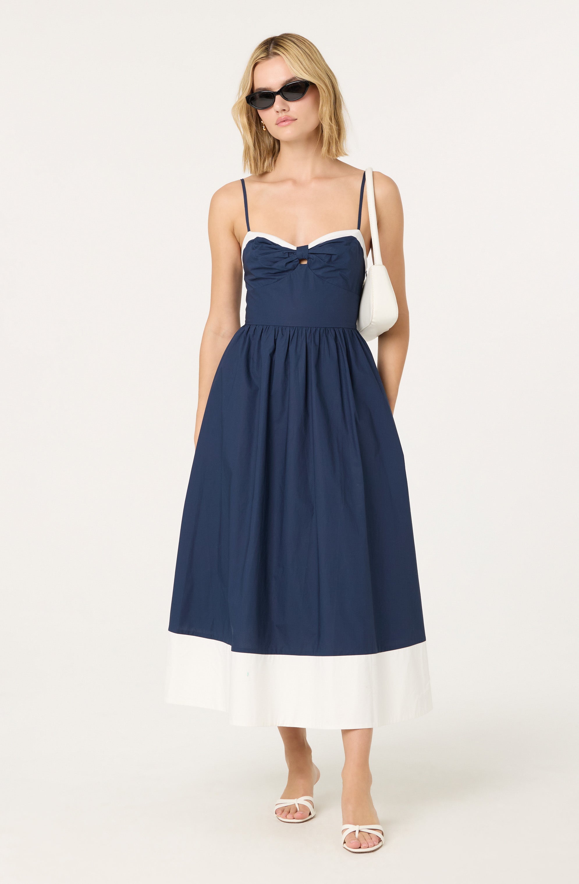 A woman is wearing a navy blue spaghetti strap midi dress with a white trim around the neckline and a bow detail at the bust. She accessorizes with black sunglasses, a small white shoulder bag, and white open-toed sandals with bows.