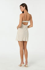 A woman is wearing a sleeveless, spaghetti strap dress that features a cream-colored fabric with a brown polka dot pattern. The back of the dress has a cutout section that exposes part of her back and is accented with a ruffled trim along the top edge. She is also wearing white high-heeled shoes and gold hoop earrings.
