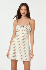 A young woman is wearing a cream-colored dress with brown polka dots. The dress has thin spaghetti straps and features a small decorative bow with a gold element at the center of the bust. The dress is fitted at the waist and flares out slightly towards the hem, creating an A-line silhouette.
