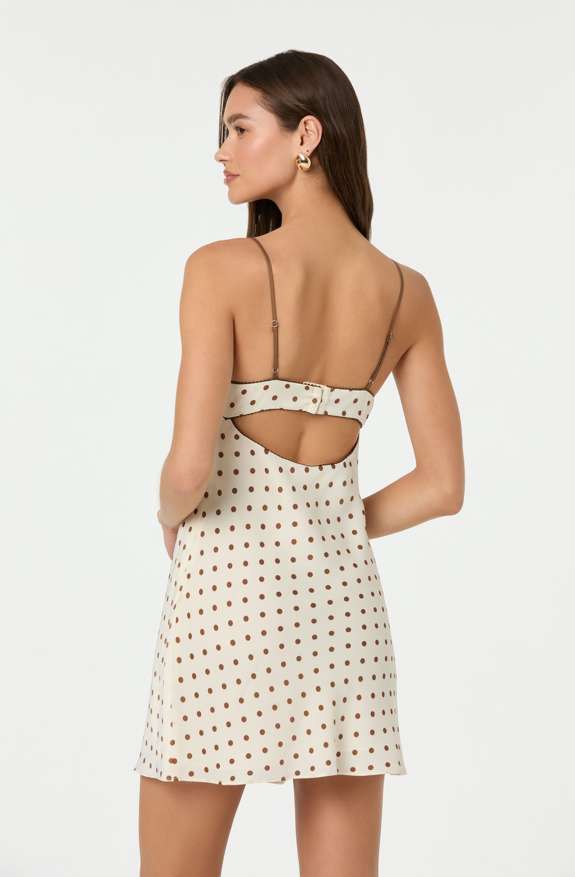 A woman is wearing a sleeveless, cream-colored dress with brown polka dots. The dress has thin spaghetti straps and features a cut-out design on the back with a horizontal strap across the upper back.