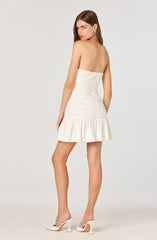 A woman is wearing a strapless, form-fitting cream-colored dress with ruching details throughout and a ruffled hem that falls above the knee. She is also wearing white high-heeled shoes with ankle straps.