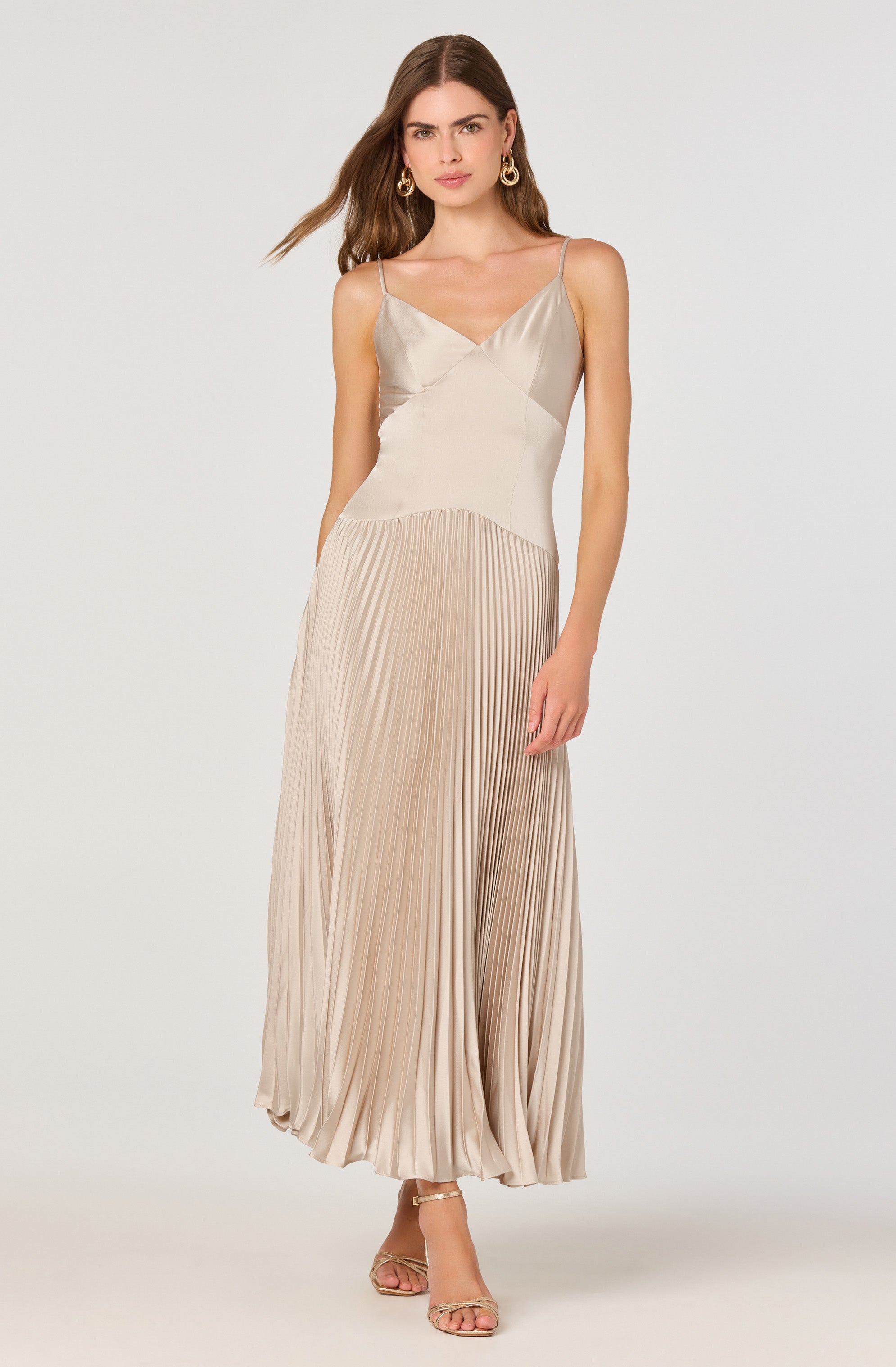 A woman is wearing a sleeveless, beige-colored dress with thin spaghetti straps. The top part of the dress has a smooth, satin-like finish with a V-neckline, while the skirt features a pleated design that creates a flowing and elegant silhouette. The dress is floor-length, and she is wearing open-toe heels.