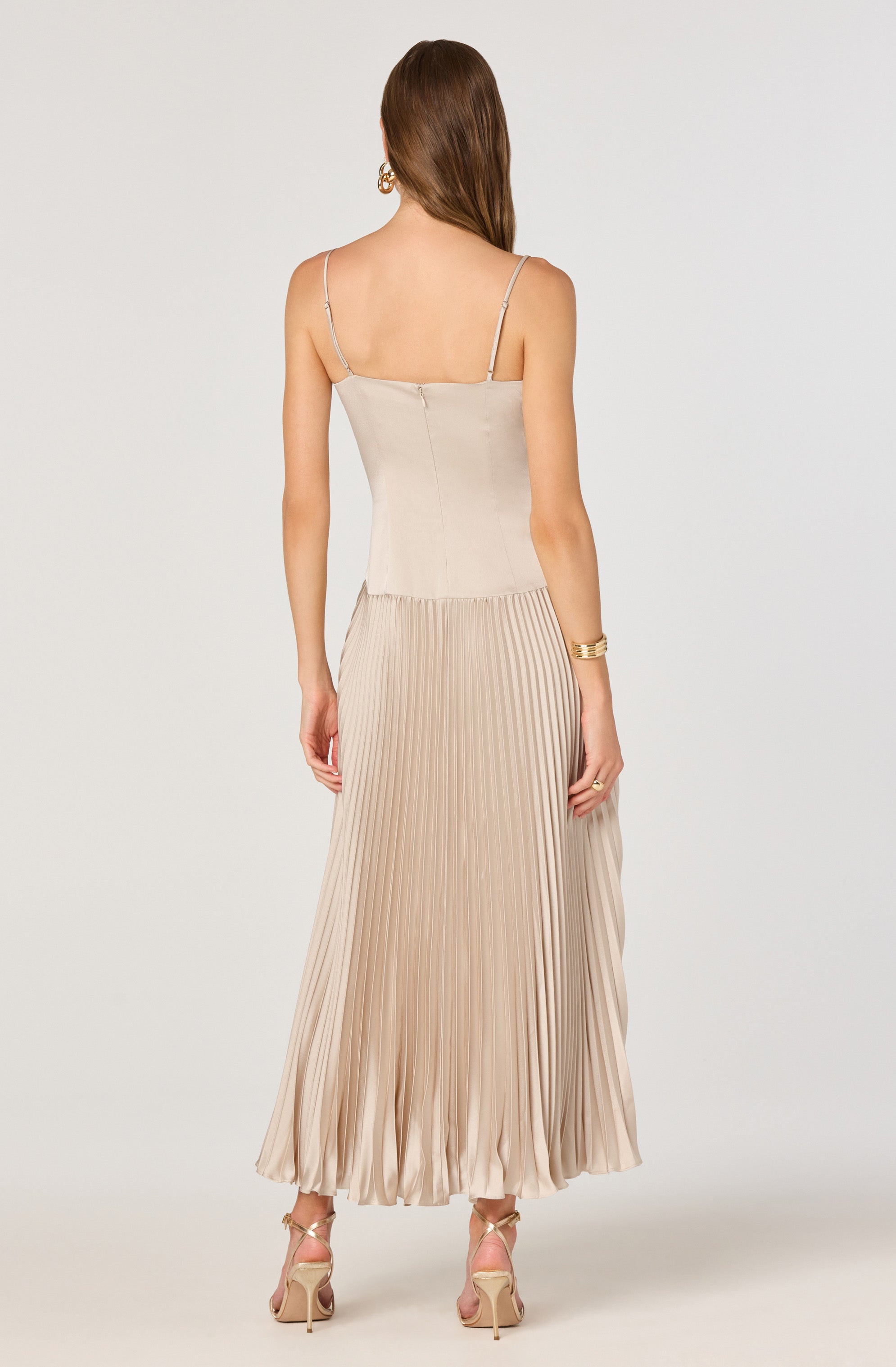The woman is wearing a beige sleeveless top with thin spaghetti straps and a high-waisted, pleated, satin maxi skirt in a matching shade. She also has on high-heeled sandals with ankle straps, large hoop earrings, and a gold bangle bracelet on her left wrist.
