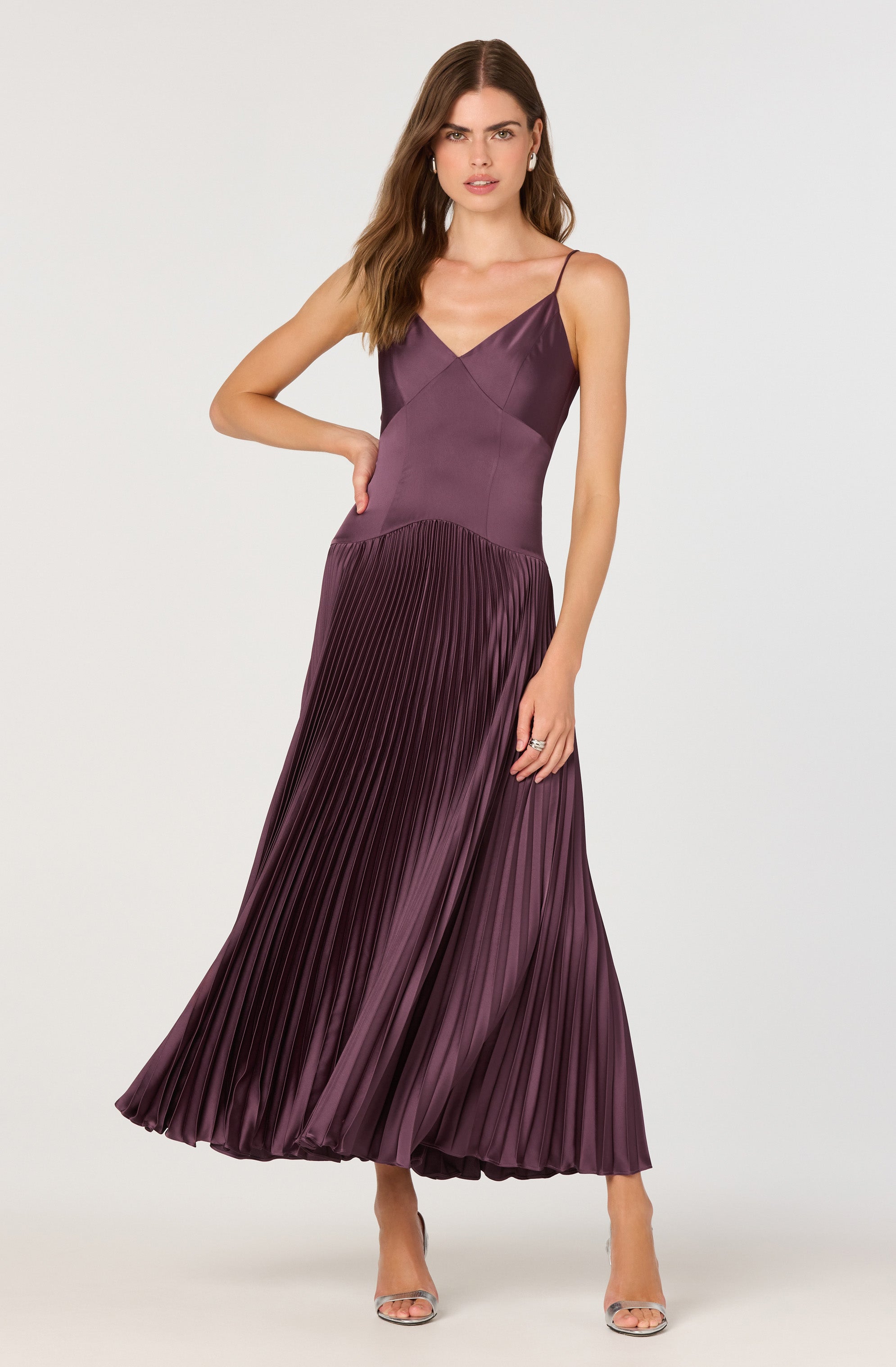 A woman is wearing a sleeveless, floor-length gown with thin spaghetti straps. The dress has a fitted bodice and a full, pleated skirt made of shiny satin fabric in a rich, deep purple color.