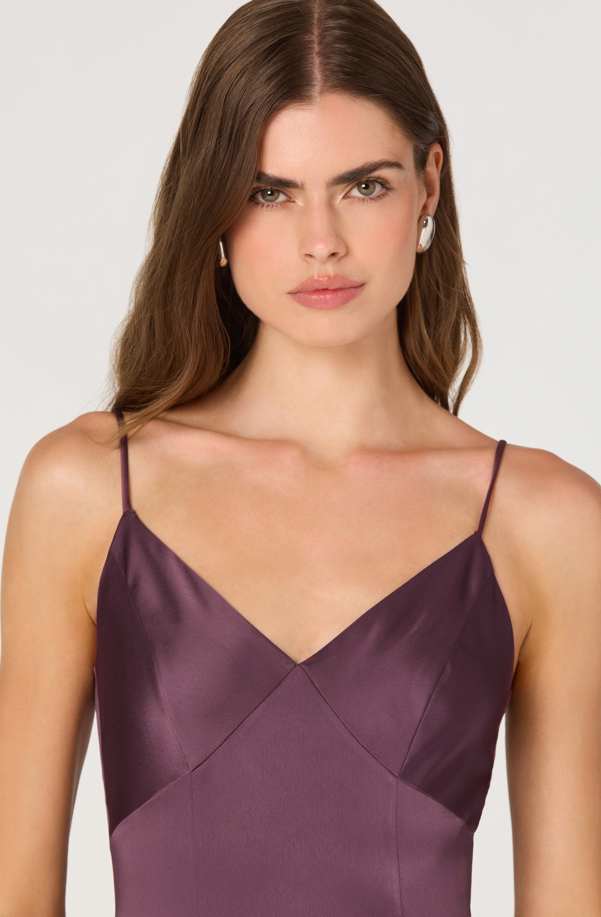 A woman is wearing a sleeveless dress with thin spaghetti straps. The dress has a satin finish and features a color-block design with shades of dark purple and plum, creating a diagonal pattern across the bodice.