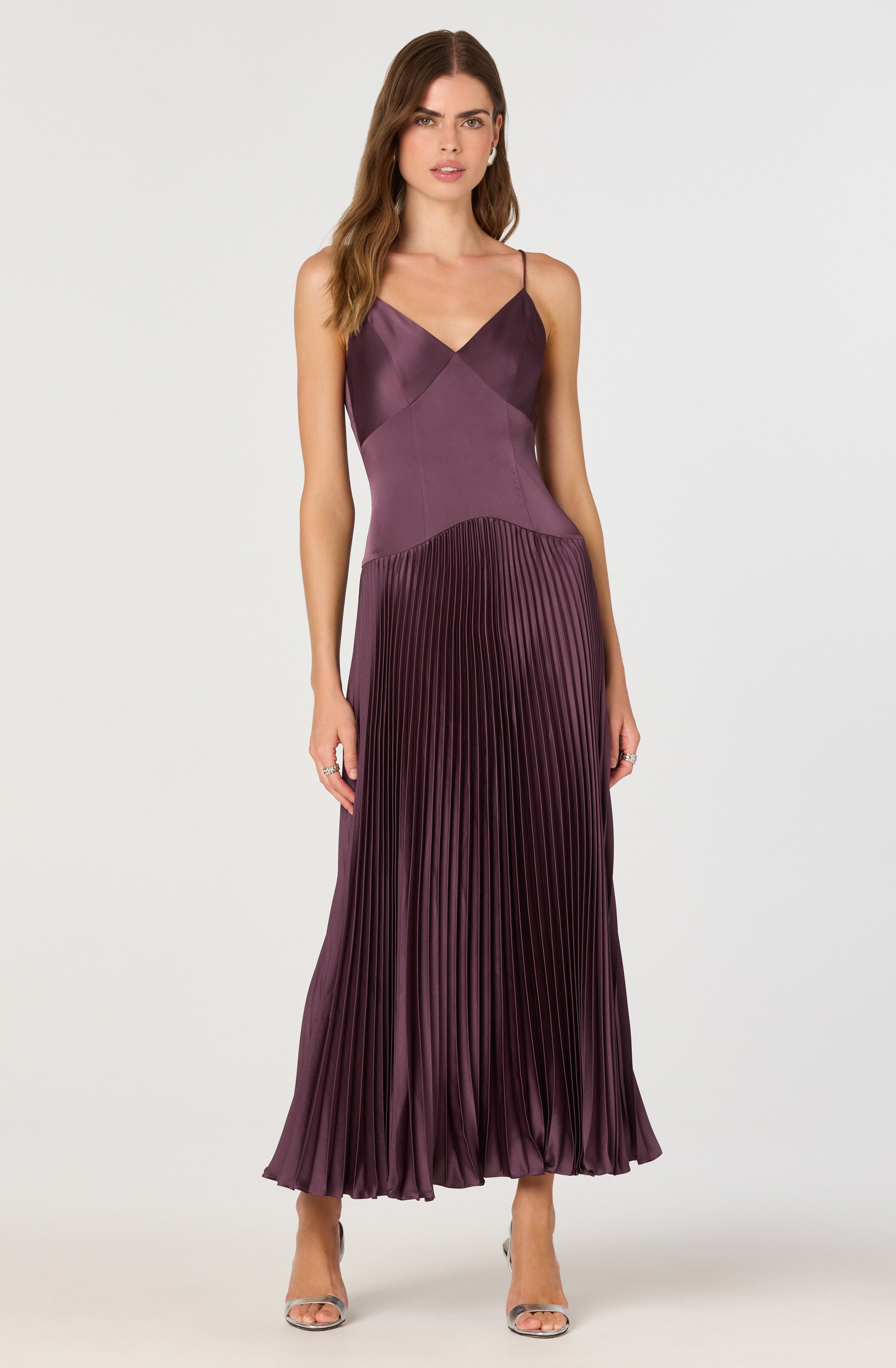 A woman is wearing a sleeveless, floor-length gown with spaghetti straps. The top part of the dress features a fitted, satin-like bodice in a deep plum color, with diagonal seam details. The skirt is made of a pleated fabric in a matching purple shade, flowing to the floor with evenly spaced vertical pleats. The outfit is completed with open-toe high-heeled shoes.