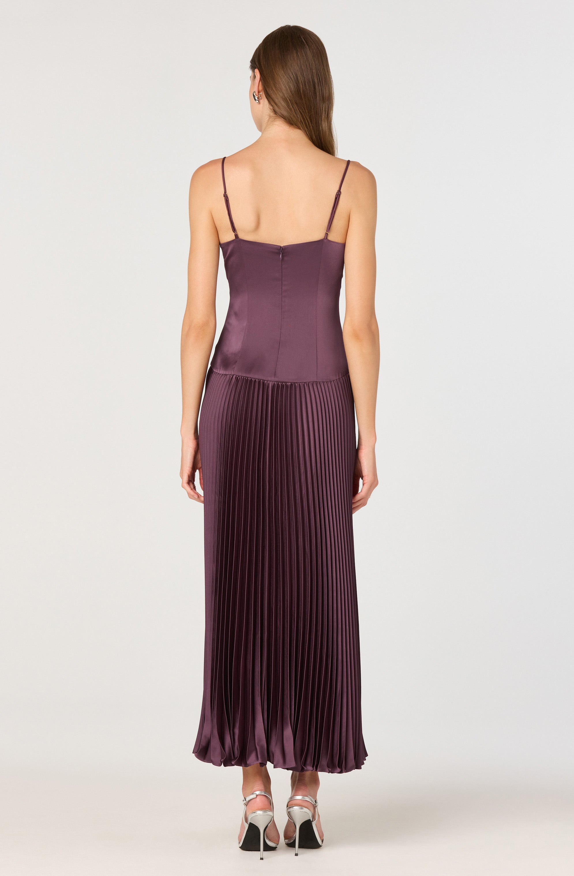 A woman is wearing a sleeveless, purple satin dress with thin spaghetti straps. The top part of the dress is fitted, while the skirt features a pleated design that extends to the floor, creating a flowing silhouette. The fabric has a shiny finish, adding an elegant touch. She is also wearing high-heeled shoes with straps.