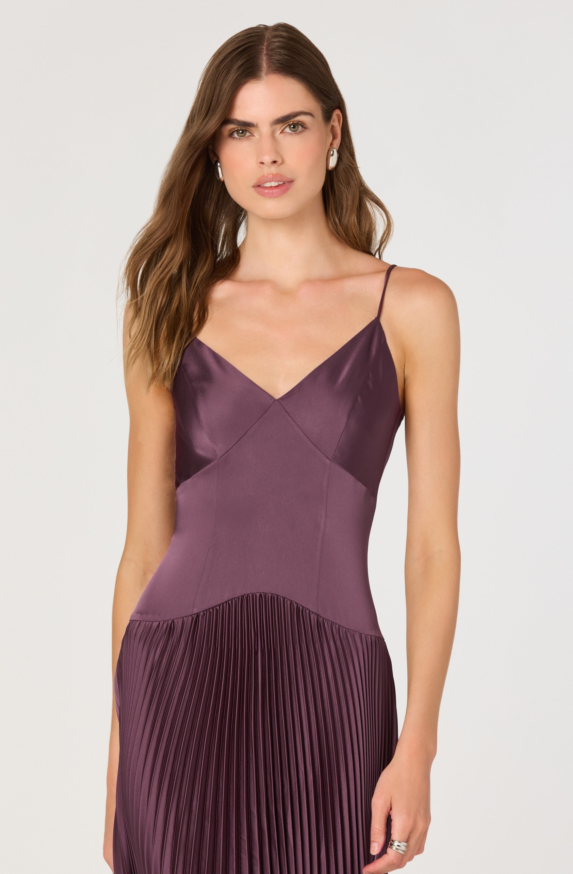 A woman is wearing a sleeveless dress with thin spaghetti straps. The top part of the dress is made of a shiny, satin-like fabric in a deep purple color with a V-neckline. The midsection has a matte, smooth fabric in a lighter purple shade. The skirt features a pleated design with vertical pleats in a darker purple hue. She is also wearing stud earrings and a ring on her right hand.