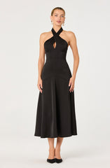 A woman is wearing a sleeveless black halter dress with a crisscross neckline that forms a keyhole cutout at the chest. The dress cinches at the waist and flows into a wide, A-line skirt that reaches mid-calf. She is accessorized with large pearl earrings and black shoes with bows.
