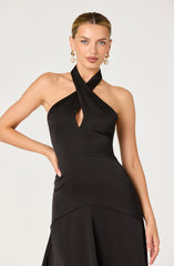 A woman is wearing a sleeveless black dress with a halter neckline that features a twisted fabric detail at the neck and a small keyhole cutout at the chest. The dress has a fitted waist and a flowing skirt. She is accessorized with large pearl drop earrings.