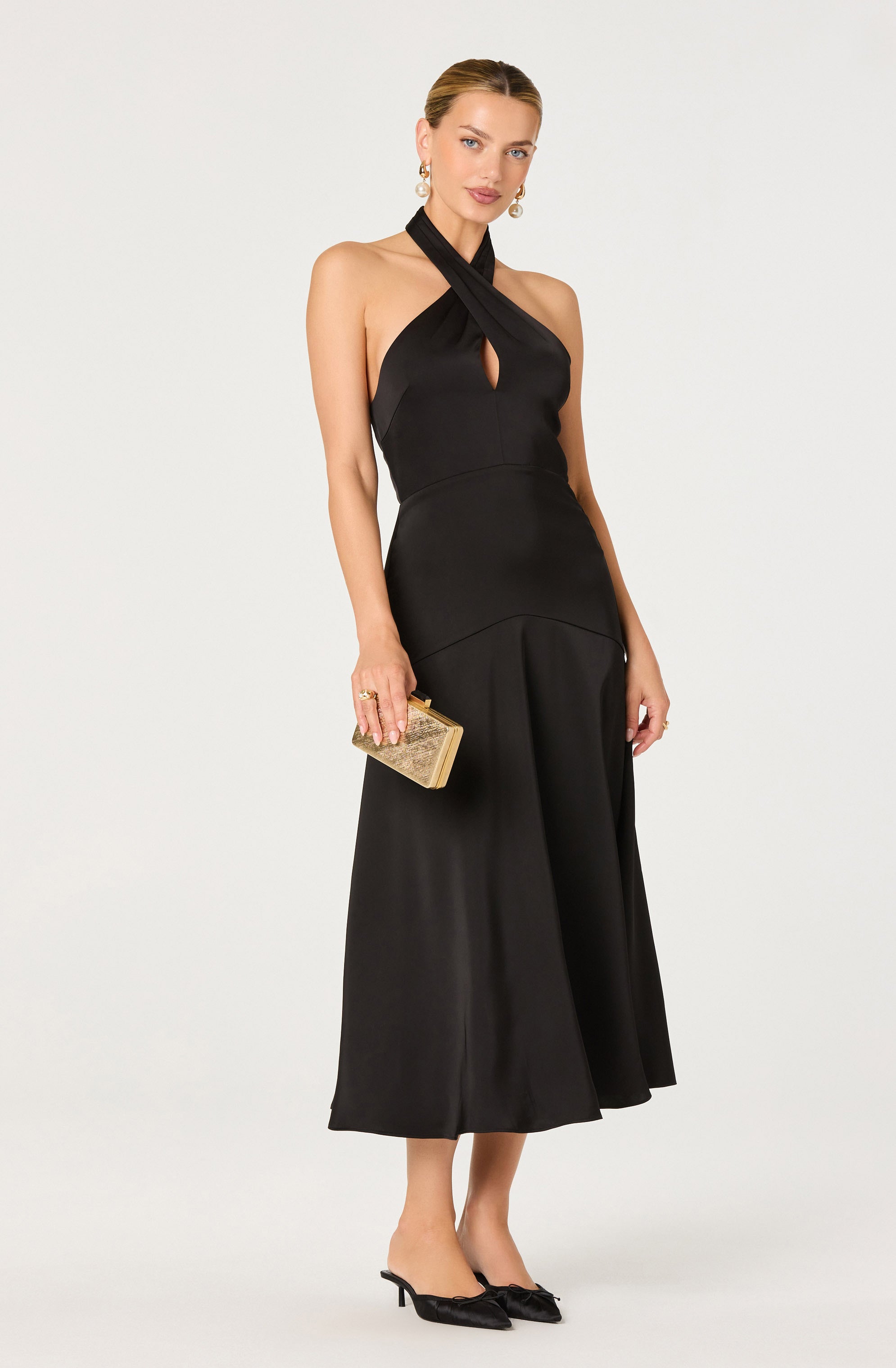 A woman is wearing a sleeveless, black halter-neck dress with a keyhole cutout at the chest. The dress has a fitted bodice and a flared, midi-length skirt. She is holding a small, gold, embellished clutch purse and is wearing black high-heeled shoes with bows on the toes. She has pearl earrings and her hair is styled in a sleek, pulled-back fashion.