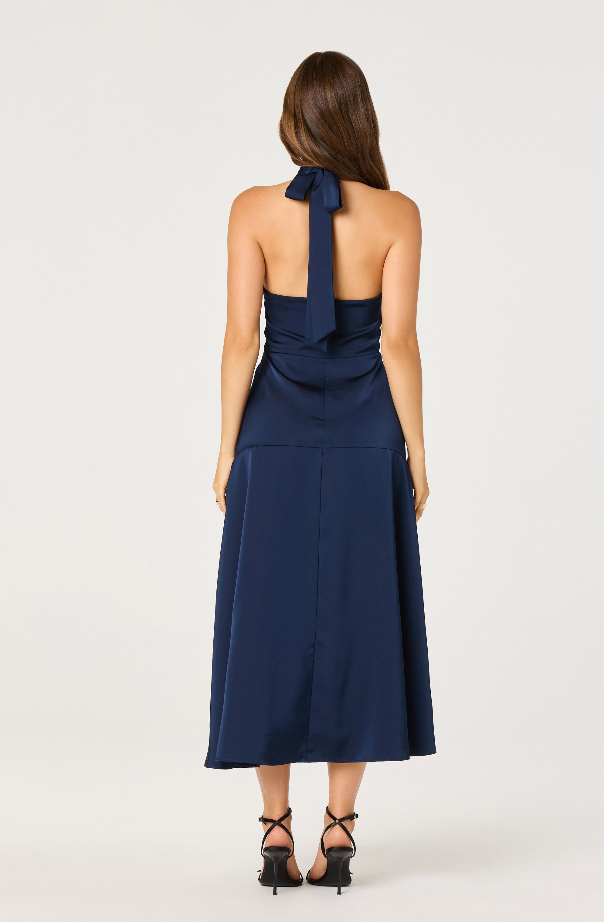 A woman is wearing a strapless navy blue dress with a fitted bodice and a wide waistband. The dress features a halter-style neckline with a large bow at the back of her neck, and the skirt is long, flowing, and reaches mid-calf. She is also wearing black high-heeled sandals with thin ankle straps.