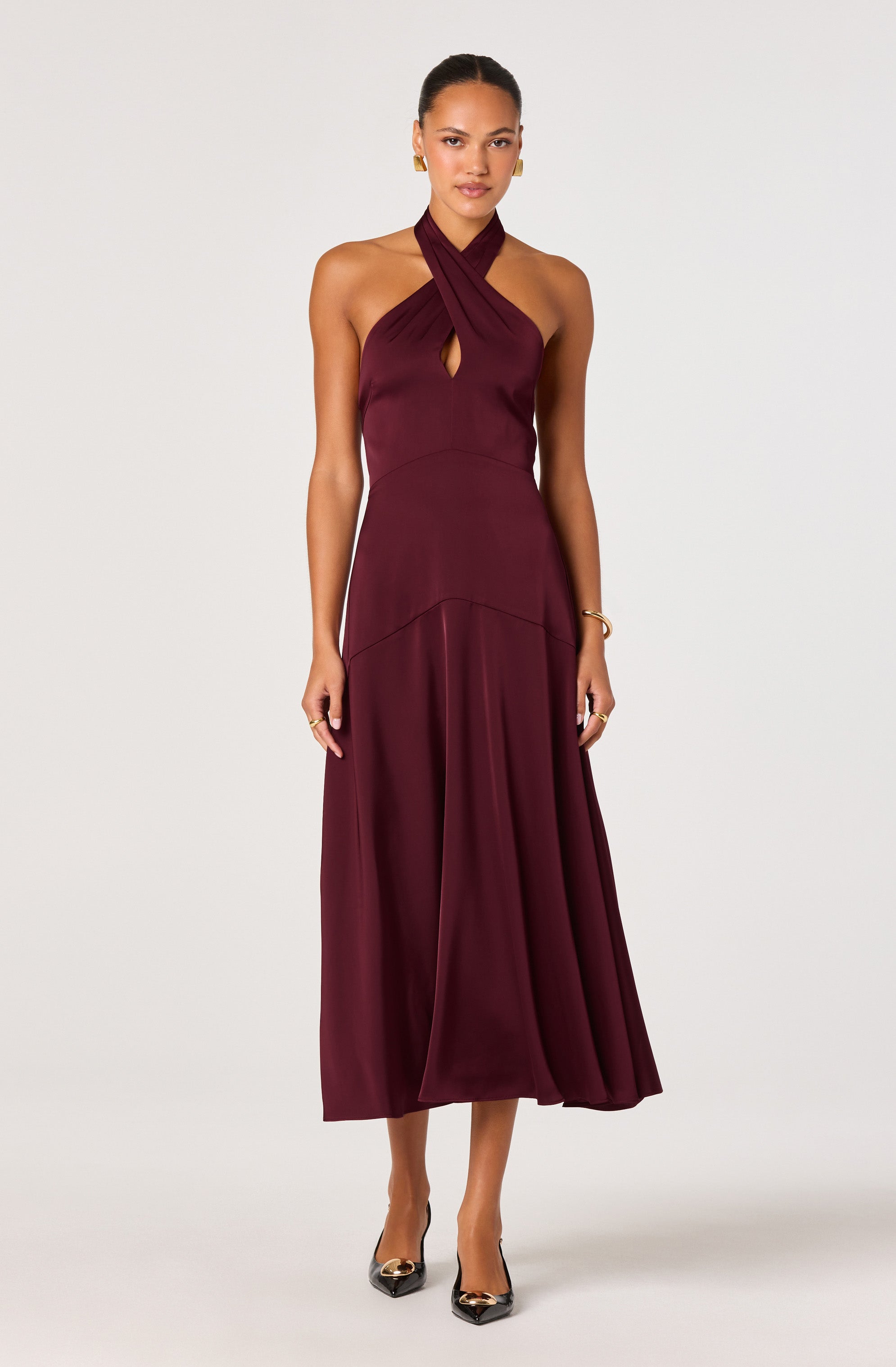 A woman is wearing a sleeveless, deep burgundy halter dress with a pleated skirt. The neckline features a crisscross design with a keyhole cutout at the chest. She is accessorized with gold earrings, a bracelet, and rings, and is wearing black high-heeled shoes with a metallic embellishment on the toe.