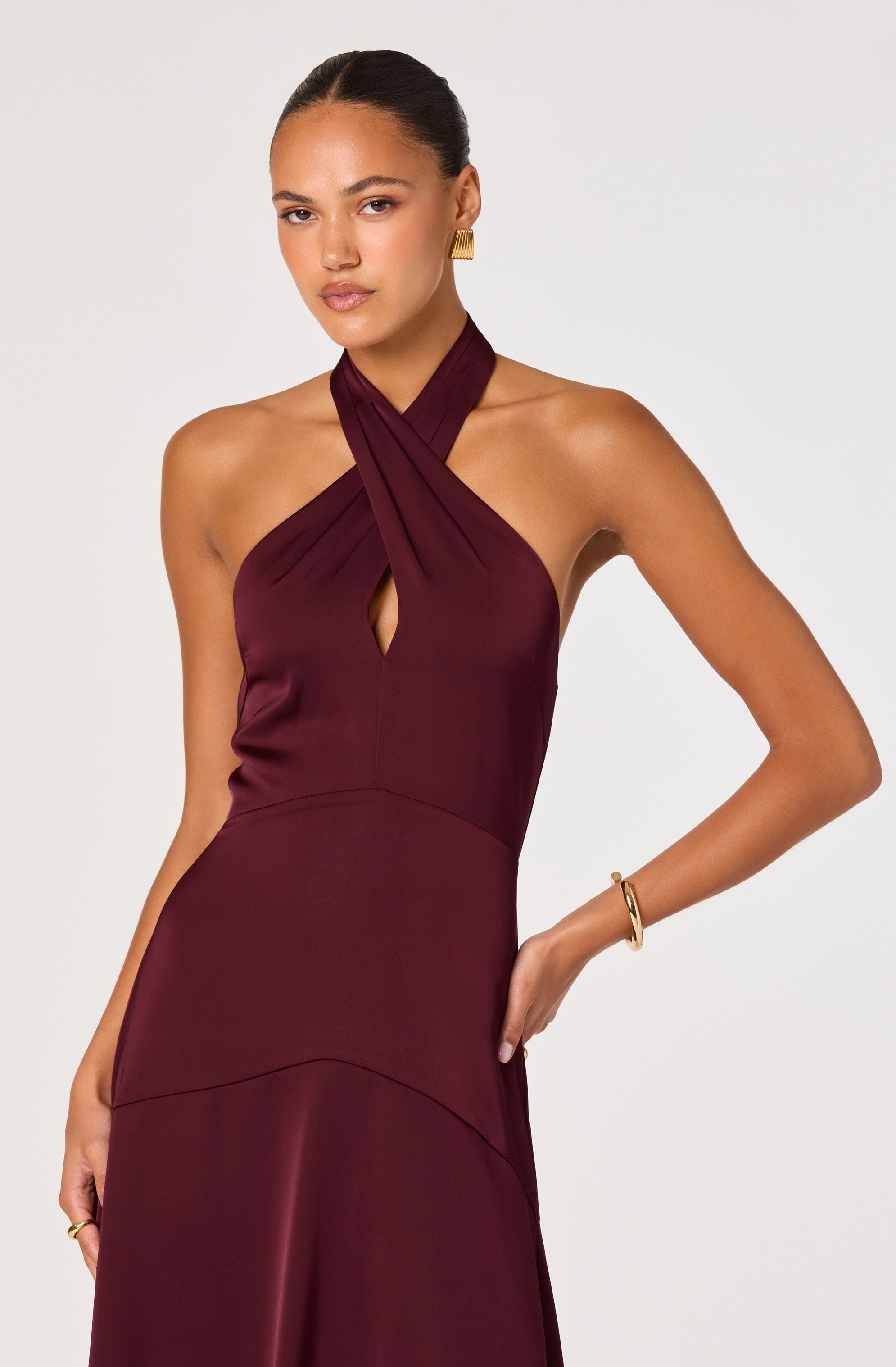 A woman is wearing a sleeveless, floor-length burgundy dress with a halter neckline that features twisted fabric details crossing at the neck and forming a keyhole opening at the chest. The dress is fitted at the waist and has a smooth, satin-like finish.