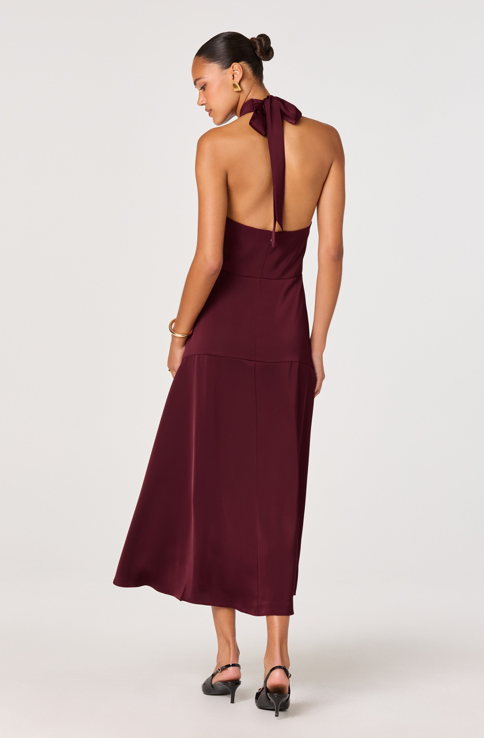 A woman is wearing a sleeveless, high-waisted, deep burgundy halter dress with a large bow tied at the back of the neck. The dress is long, reaching her ankles, with a smooth, satin-like texture. She accessorized with gold earrings and a gold bracelet, and is wearing black high-heeled sandals.