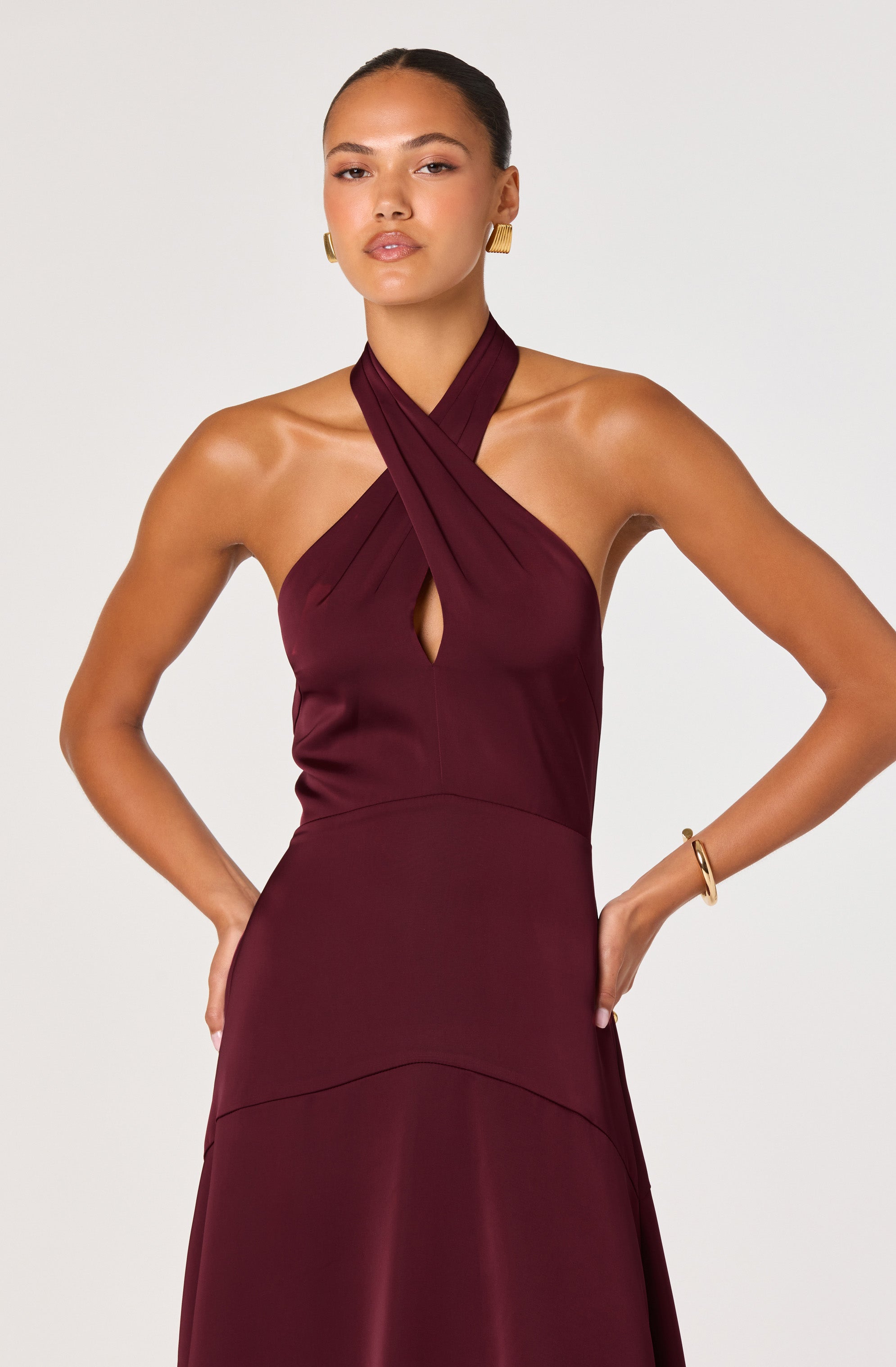 A woman is wearing a sleeveless, burgundy dress with a halter neckline that crosses over at the neck and features a small keyhole opening at the center. The dress is fitted at the waist and flows into an A-line skirt. She has gold earrings and a gold bracelet on her wrist.