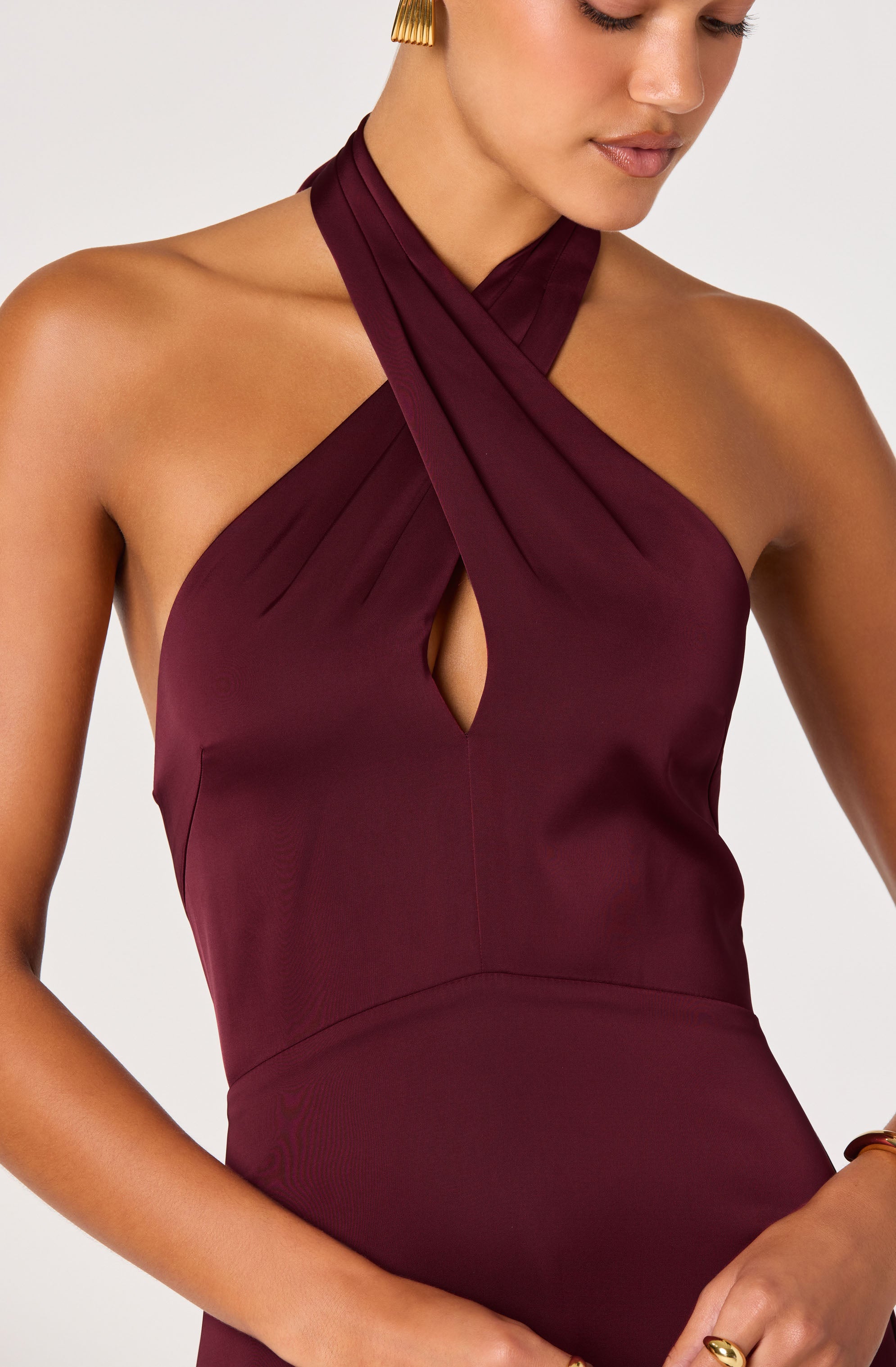 A woman wearing a sleeveless, deep burgundy dress with a halter neckline featuring layered fabric that crosses over at the neck, creating a keyhole cutout at the chest. The fabric appears smooth and slightly shiny, emphasizing elegant, draped design details.