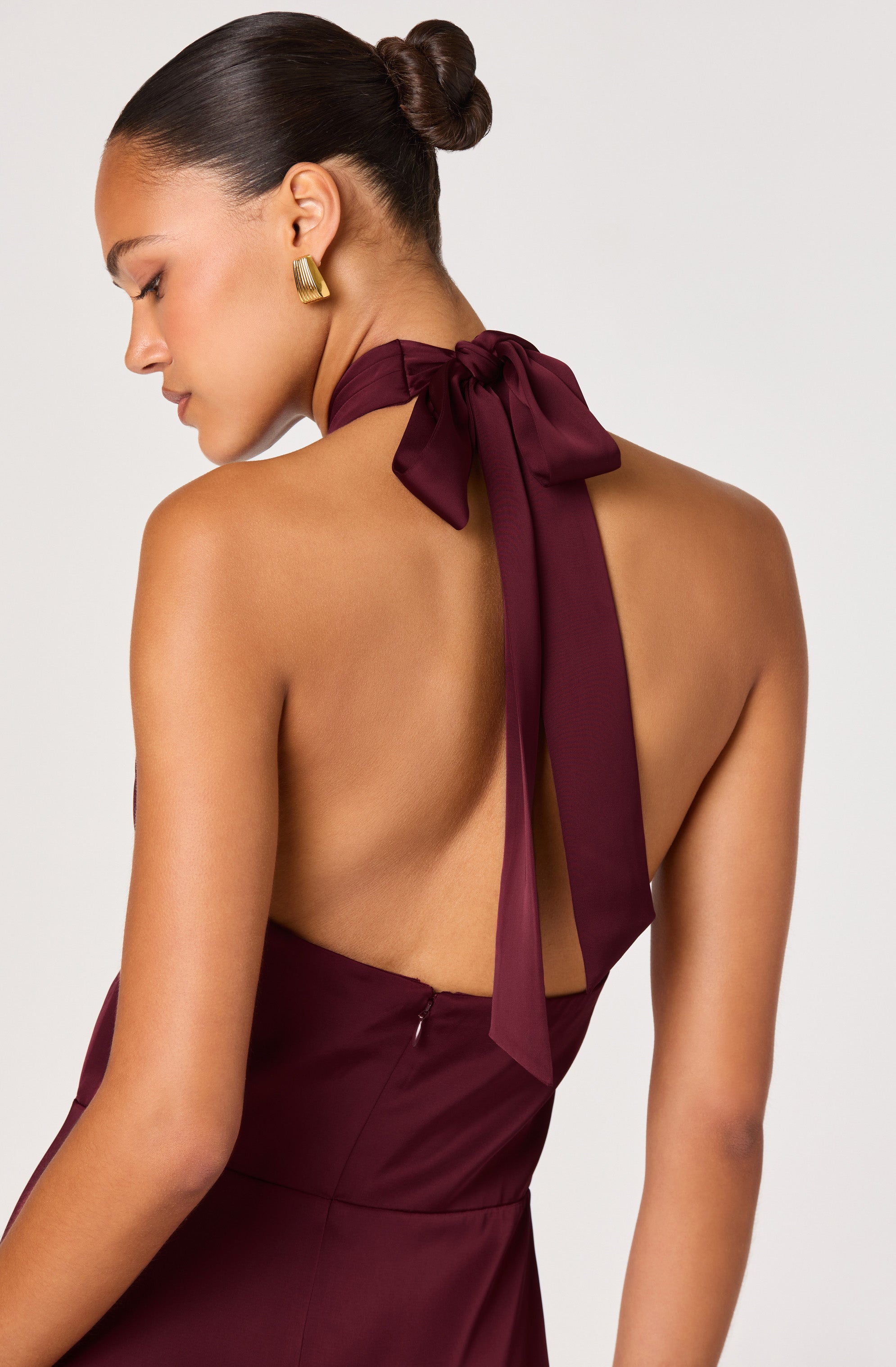 A woman is wearing a burgundy satin dress with a halter neckline tied in a large bow at the back of her neck. The dress has a fitted bodice and an open back design. She accessorized with a pair of bold, geometric gold earrings and has her hair styled in a sleek, low bun.