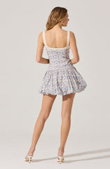 A woman is wearing a sleeveless, fitted dress with wide shoulder straps. The dress has a light-colored background with a delicate blue floral pattern, a square neckline, and a cinched waist. The skirt part of the dress is short, has a gathered, voluminous style, and ends above the knees. She is also wearing white heeled shoes.