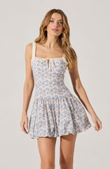 A woman is wearing a sleeveless dress with wide shoulder straps. The dress features a fitted bodice with a small bow detail at the center of the neckline and a flared, gathered skirt. The fabric has a light-colored background with a delicate blue floral print.