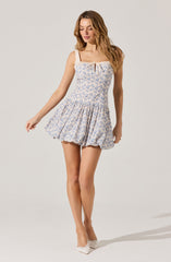A woman is wearing a sleeveless dress with wide shoulder straps and a square neckline featuring a small bow at the center. The dress has a fitted bodice and a gathered, flared skirt with a floral pattern in light blue and cream colors. She is also wearing white high-heeled shoes.