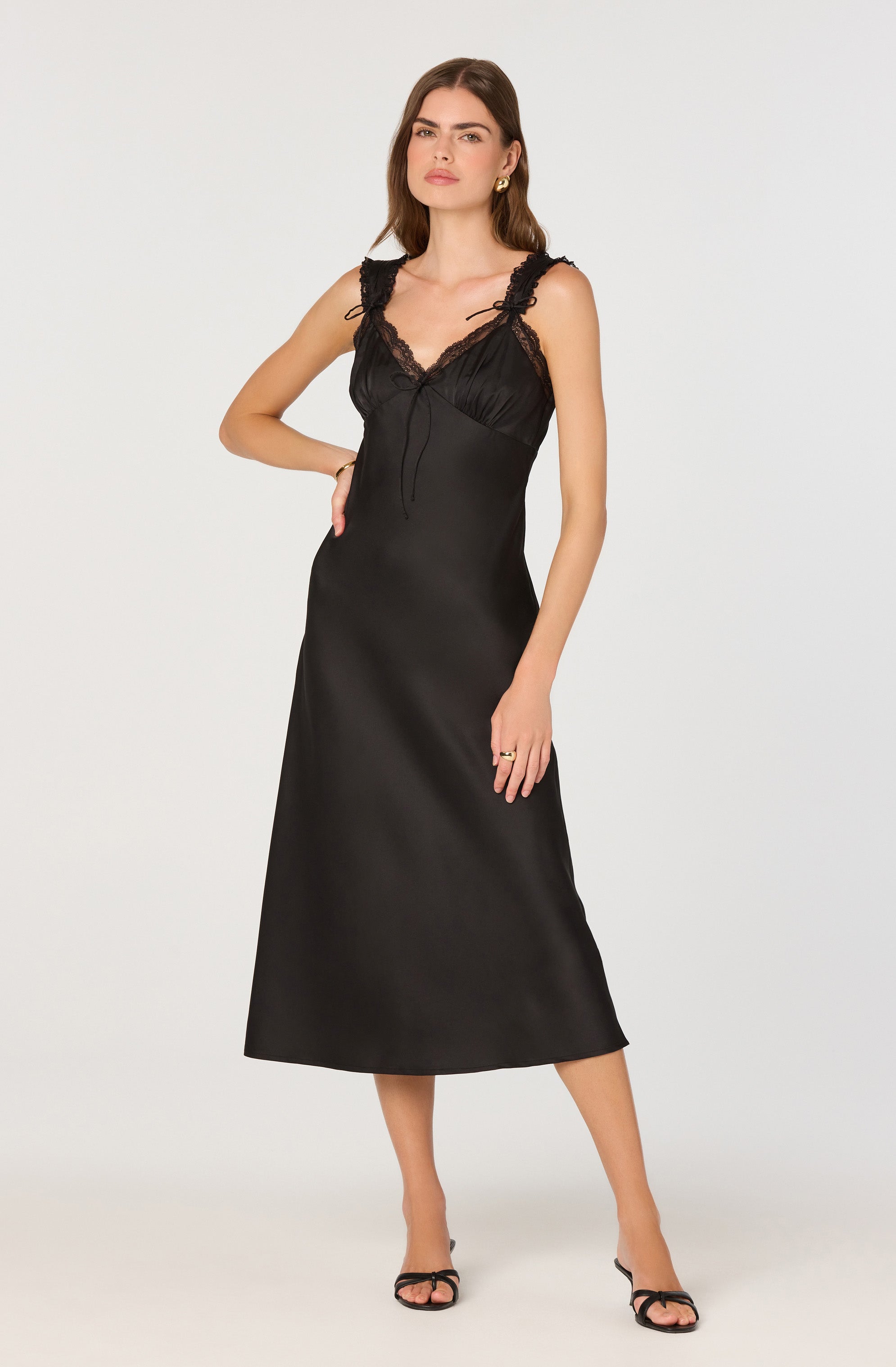 The woman is wearing a sleeveless black satin dress that features lace and ribbon details on the shoulder straps and at the neckline. The dress has a fitted bodice with lace trim and a flowy, ankle-length skirt. She is also wearing black open-toe high heel sandals.