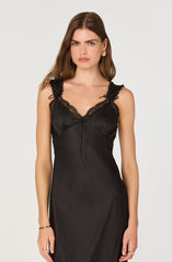 A woman is wearing a sleek black slip dress with lace detailing on the shoulder straps and neckline. The dress has a smooth, shiny fabric and a slim fit that extends below the waist. She accessorizes with gold hoop earrings, a gold bracelet, and rings on both hands.