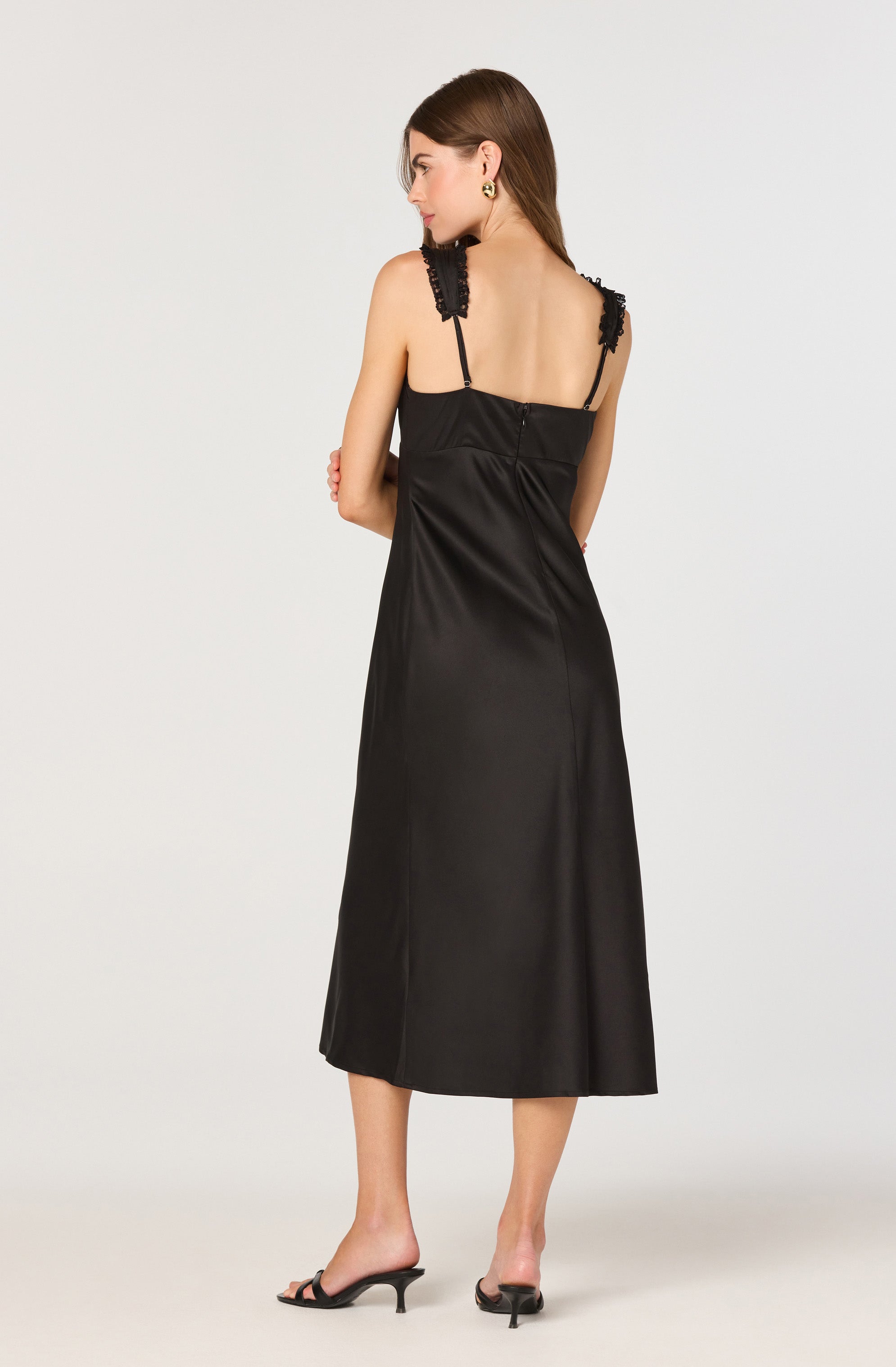 A woman is wearing a black satin sleeveless dress with lace and ribbon details on the shoulder straps. The dress has a fitted bodice and flows down to a midi length. She is also wearing black open-toe heeled sandals.